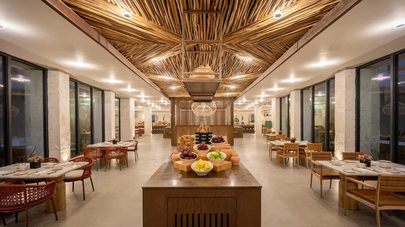 Elegant restaurant with wooden ceiling, glass walls, and neatly set tables with chairs, featuring a central dessert display with fruits.