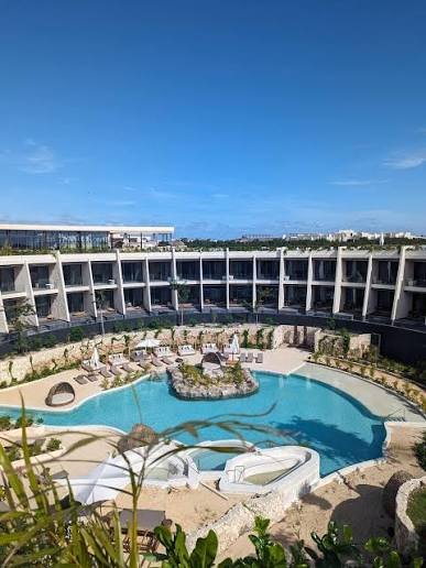 An outdoor swimming pool area with lounge chairs, surrounded by multi-story buildings and greenery, under a clear blue sky.