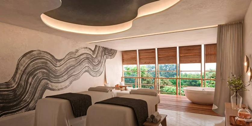 Spa room with two massage tables, a large window with wooden blinds, a freestanding bathtub, and modern decor with a mural wall and soft lighting.