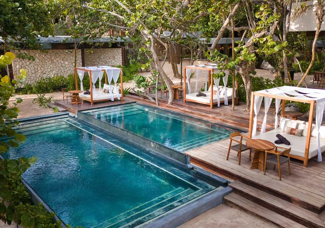 Poolside cabanas with white curtains and wooden furniture, surrounded by greenery and trees