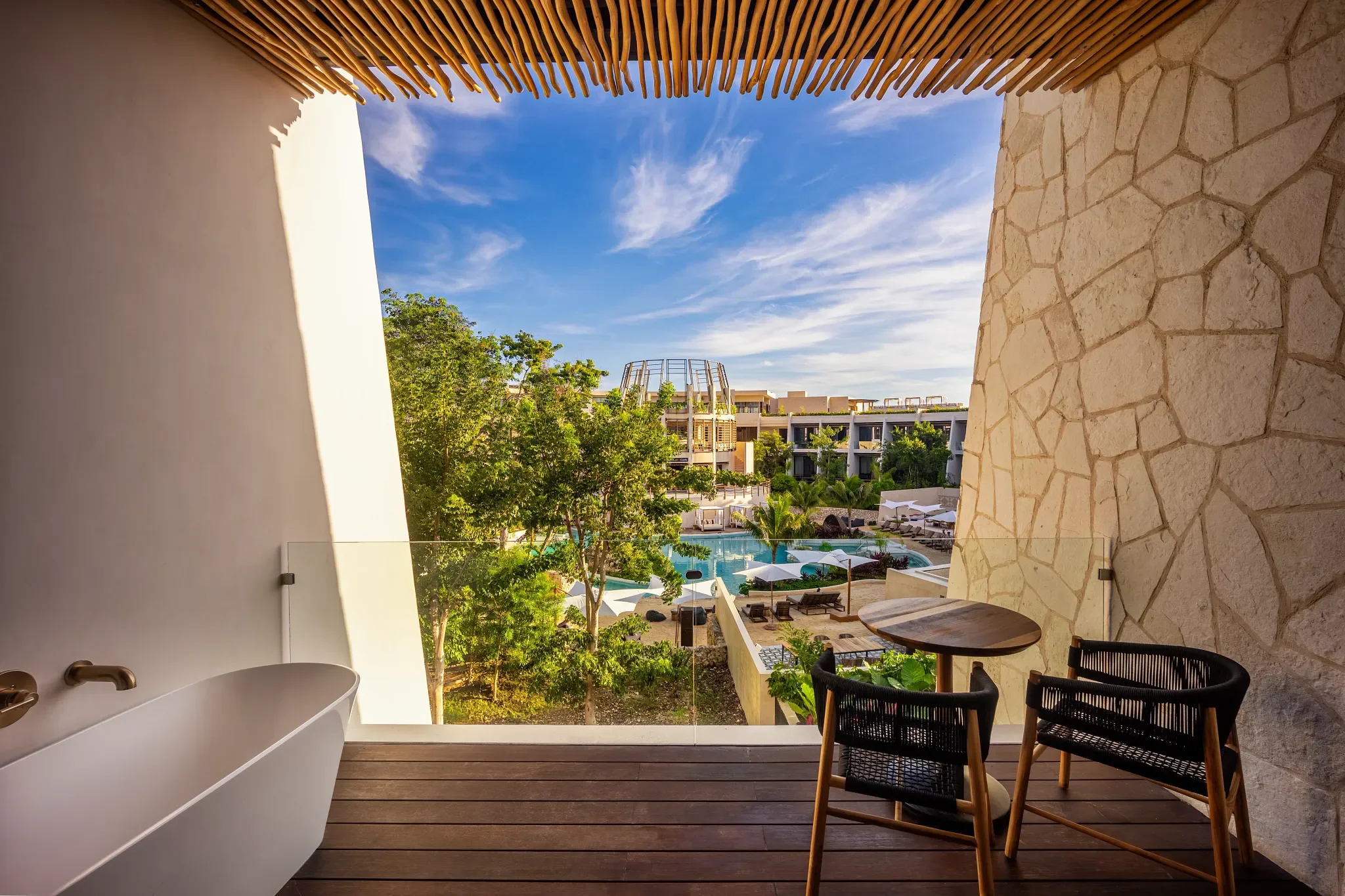 Balcony with a bathtub, two chairs, and a small table overlooking a resort with a pool, trees, and modern buildings under a blue sky.
