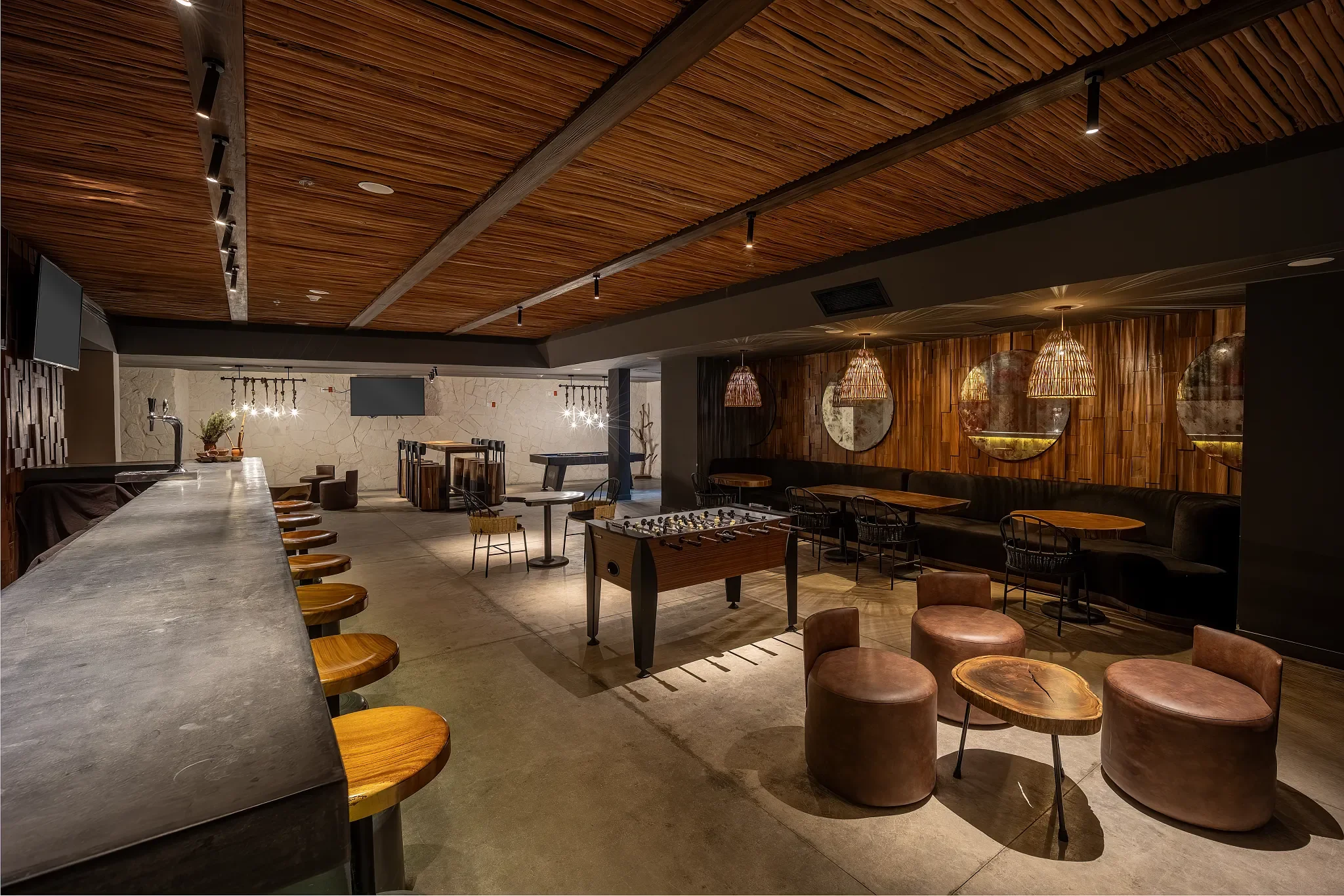A modern interior of a bar or lounge with a long concrete bar counter on the left with wooden barstools, a foosball table in the center, and seating areas with round leather stools, chairs, and tables decorated with hanging woven light fixtures and r