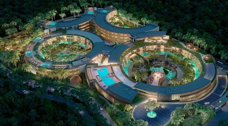 Nighttime aerial view of a large, modern resort with circular and curved structures, swimming pools, and lush greenery surrounding the property.