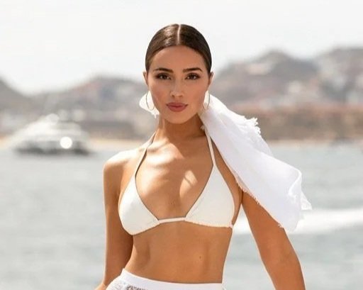 Woman in beachwear with a white bikini top and skirt, holding a white scarf, standing near water with hills in the background.