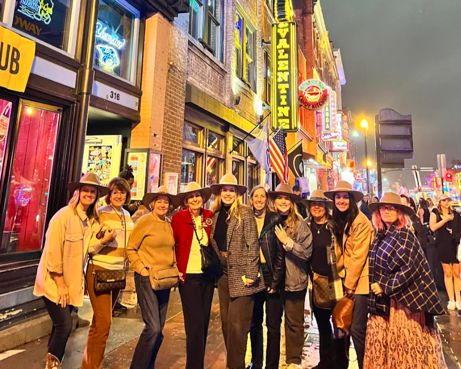A group of women wearing cowboy hats standing on a city street at night with neon signs and colorful building lights in the background.