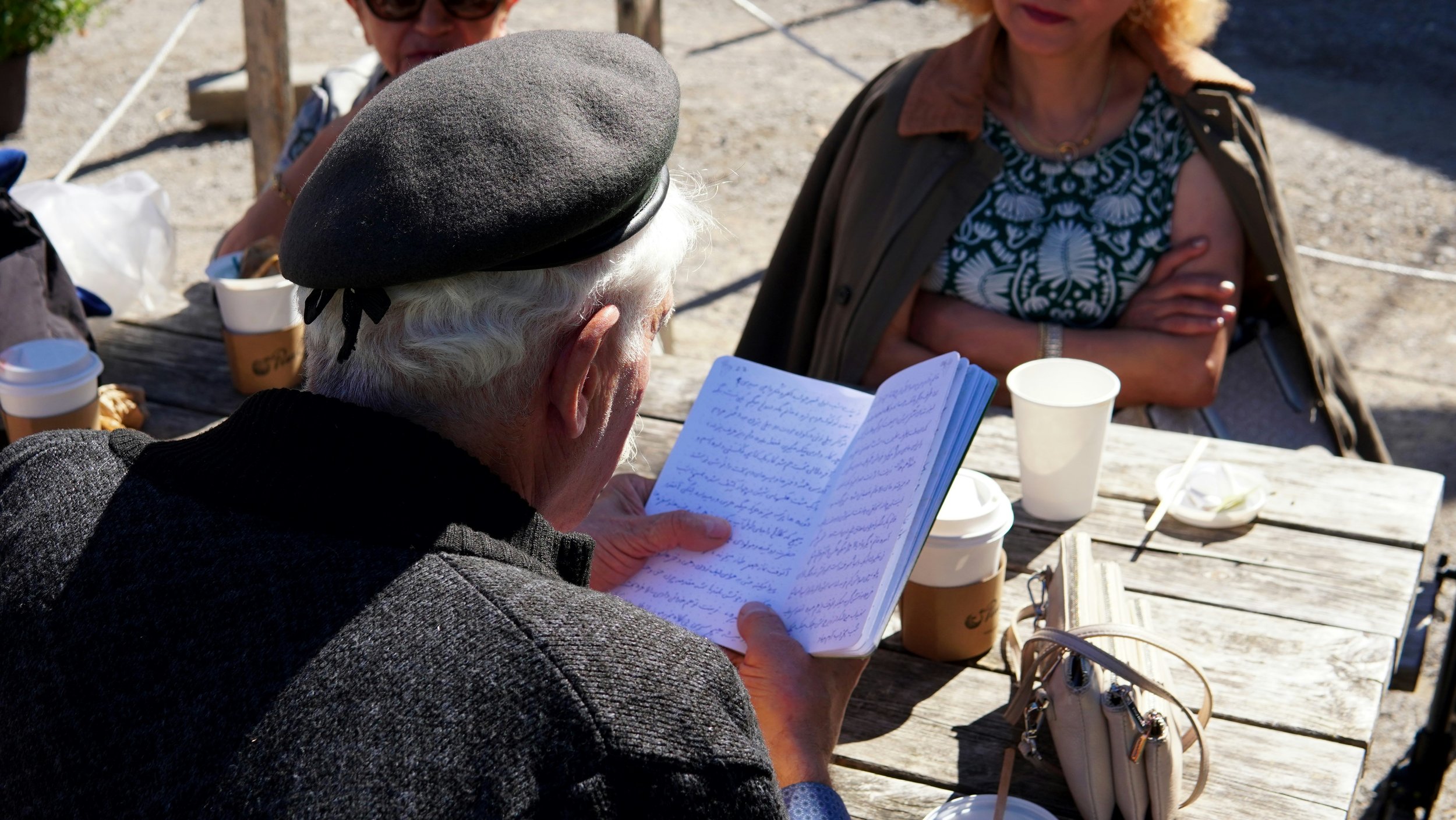 a man in a beret reading a journal at a table with people having coffee on a sunny day.
