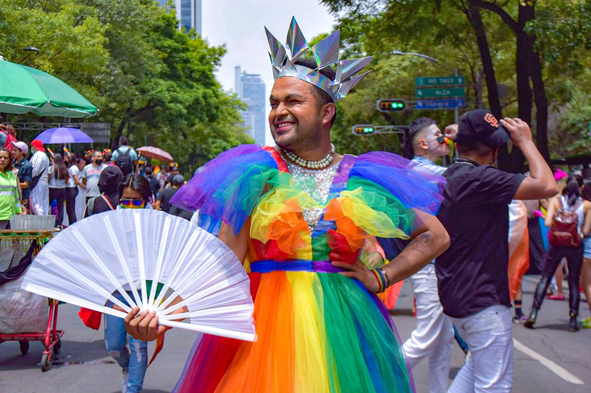 Photo at a pride march. The person is wearing a rainbow dress and a crown and holding a clacking fan while smiling