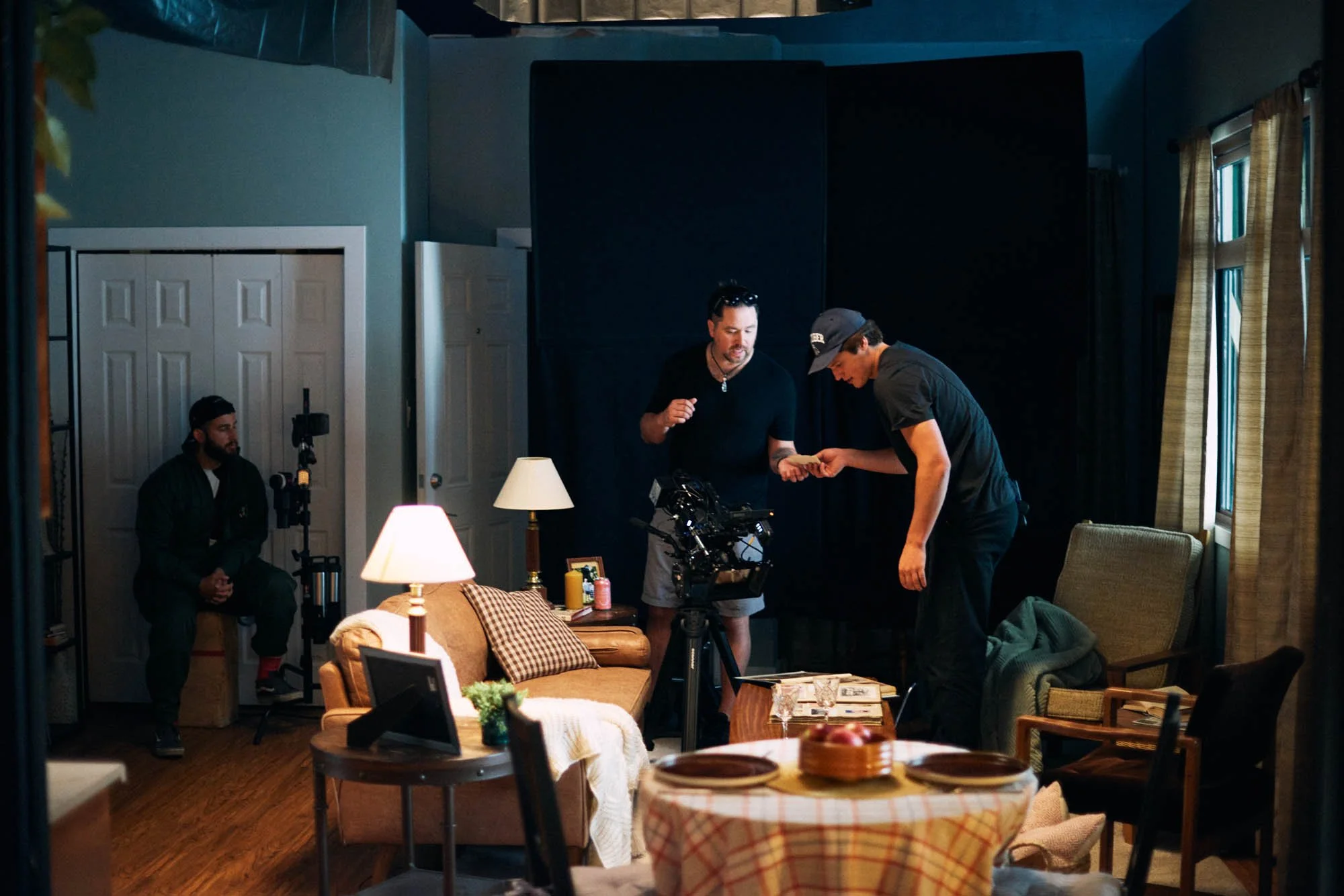 Indoor film set with crew setting up camera equipment in a living room scene, including furniture and lighting.
