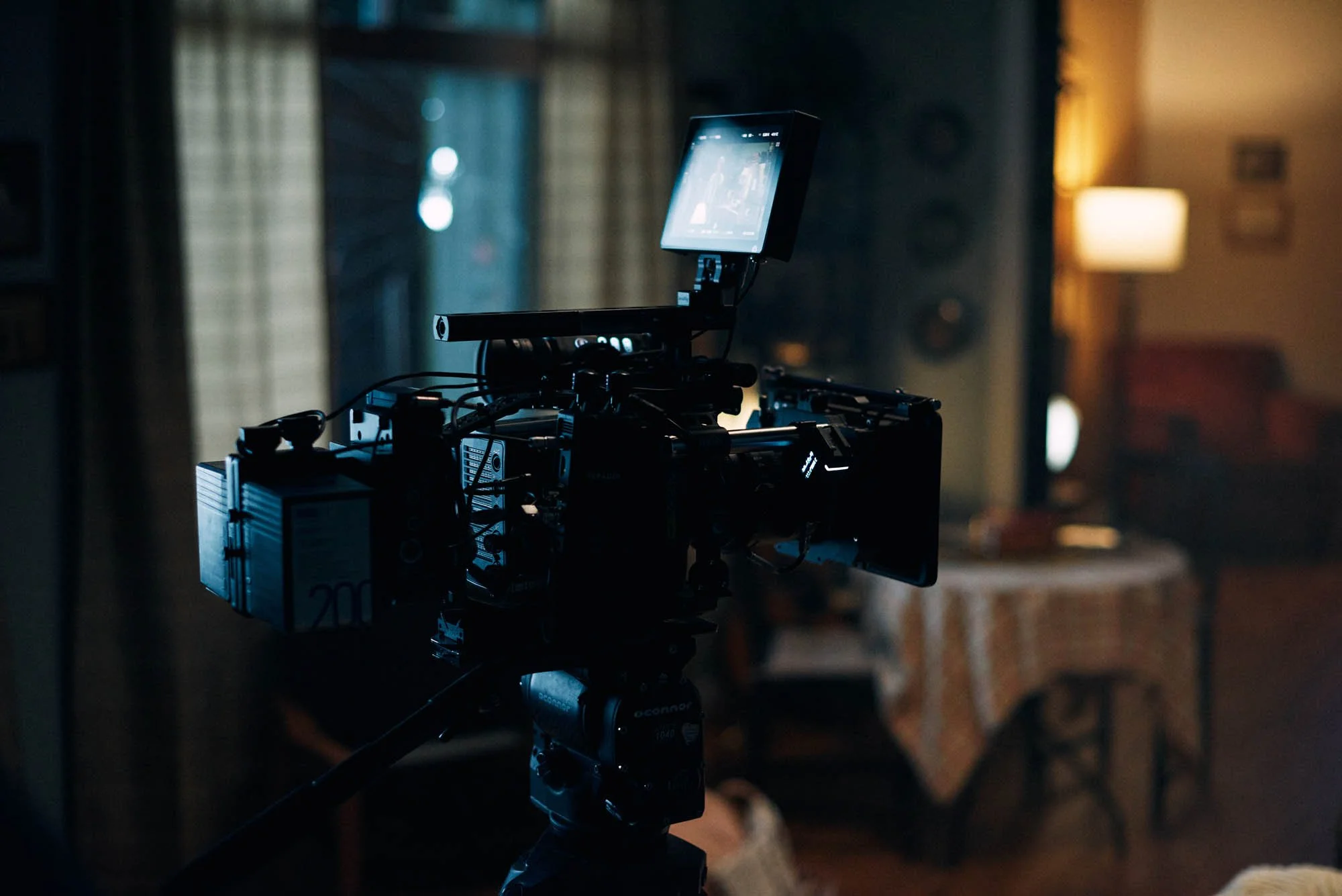 Film camera setup indoors with a monitor, blurred background with curtains and lamp.