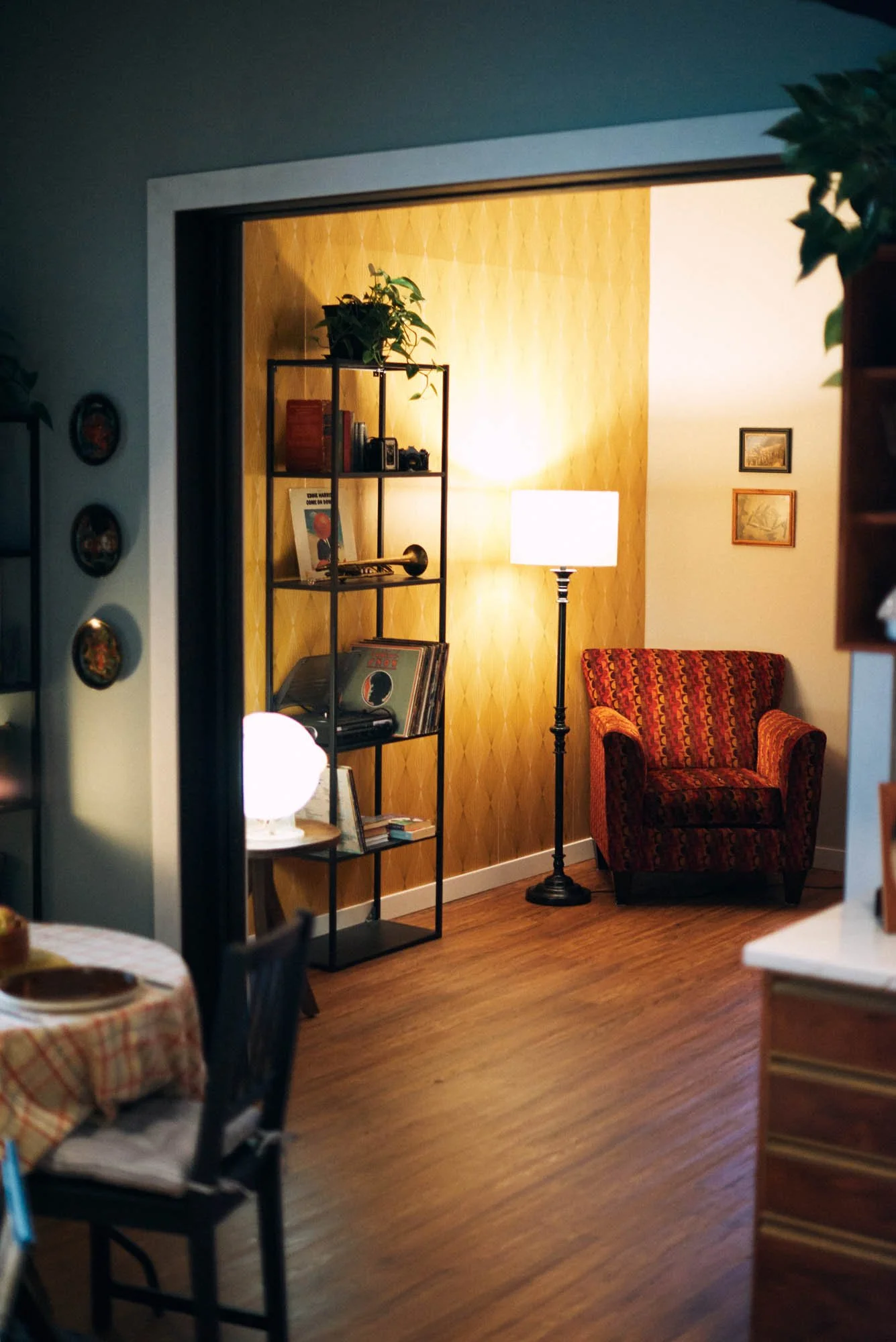 Cozy living room with an armchair, floor lamp, and bookshelf holding books and records. The walls feature yellow wallpaper and framed pictures. A dining table with a checkered tablecloth is partially visible.