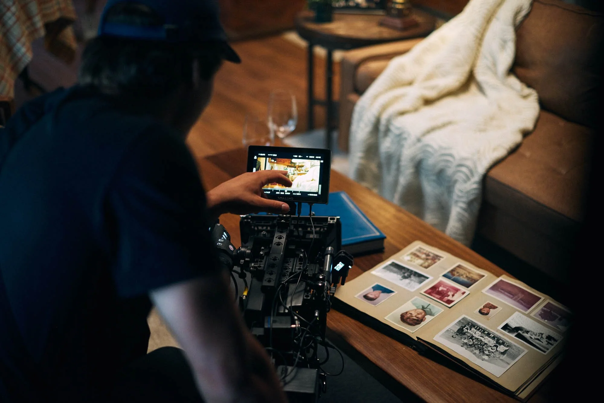Person operating a camera with a monitor filming a photo album on a table, with couches and a blanket in the background.