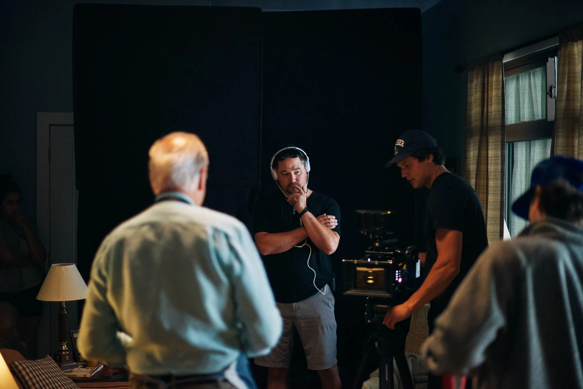 A film crew on a movie set, featuring several people, including a man wearing headphones and holding a camera. An older man faces away, and others are engaged in discussion. They're likely in a dimly lit indoor setting with equipment visible.