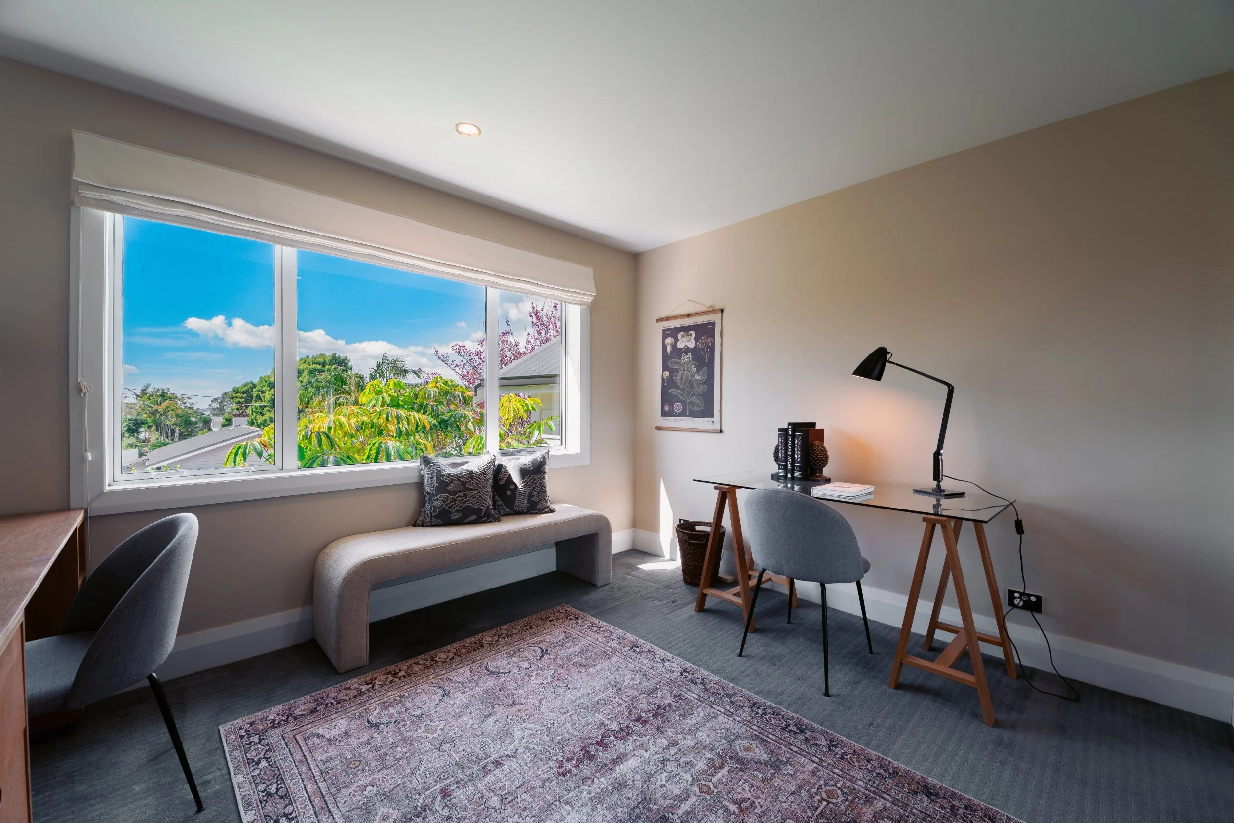A bright, cozy home office with a large window showing greenery and a blue sky. The room has a small sofa under the window with patterned cushions, a wooden desk with a gray chair, a black desk lamp, books, and artwork on the walls, all on a patterne