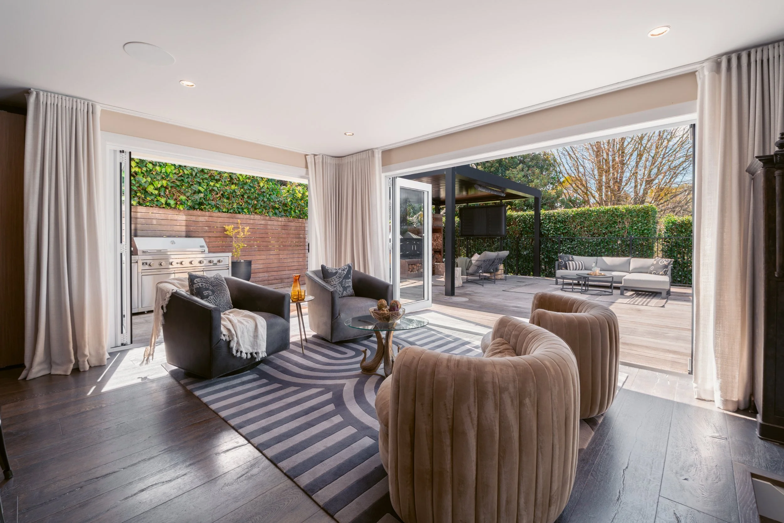 Living room with large sliding glass doors leading to an outdoor patio, featuring outdoor seating, a grill, surrounded by greenery and trees.