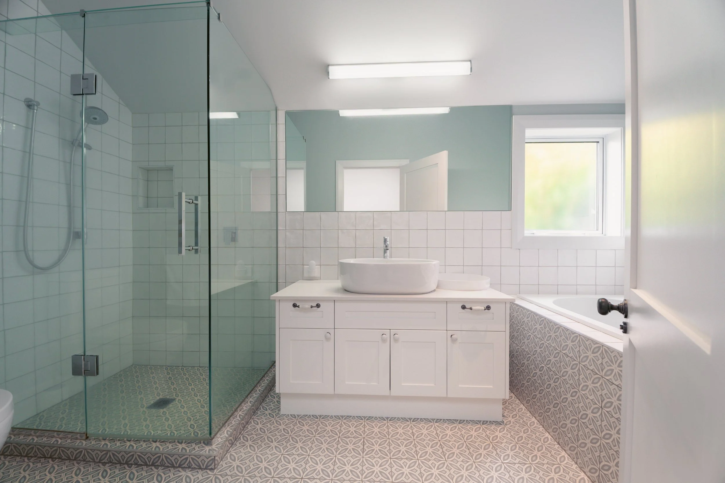 A modern bathroom with a glass shower enclosure, a white vanity with a vessel sink, a large mirror, a bathtub, and a window letting in natural light.