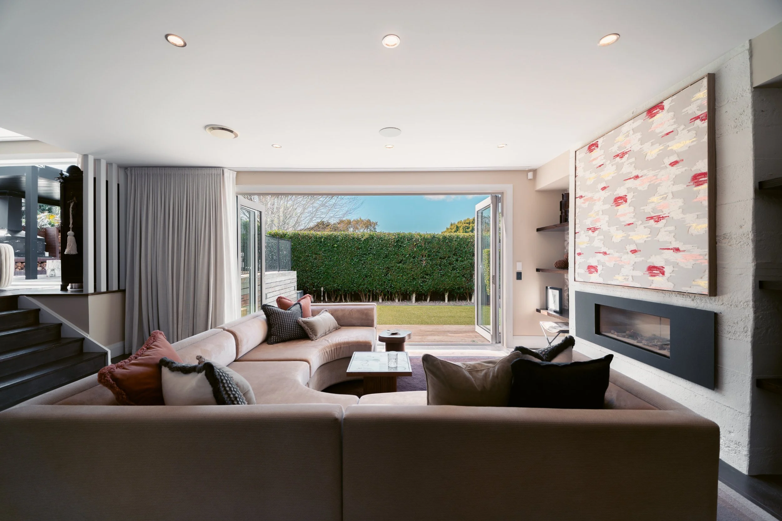 Living room with large beige sectional sofa, throw pillows, and an open sliding door leading to a backyard with green hedge and trees.