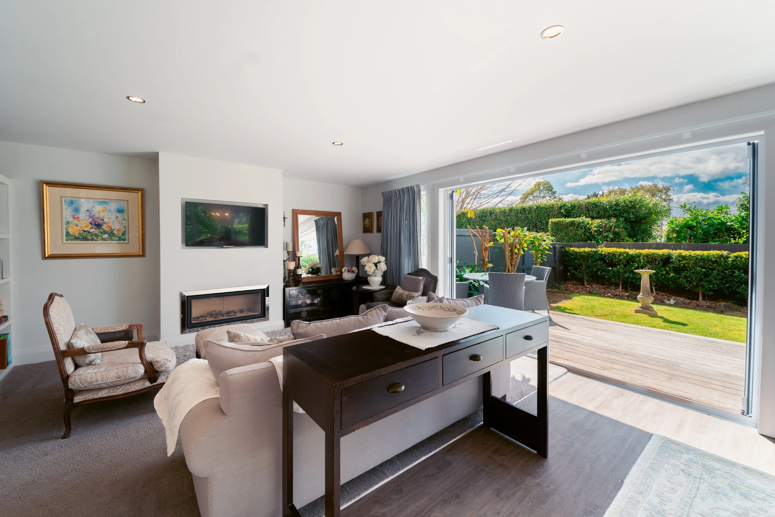 Living room with large open sliding door showing outdoor patio with garden, sky, and trees.