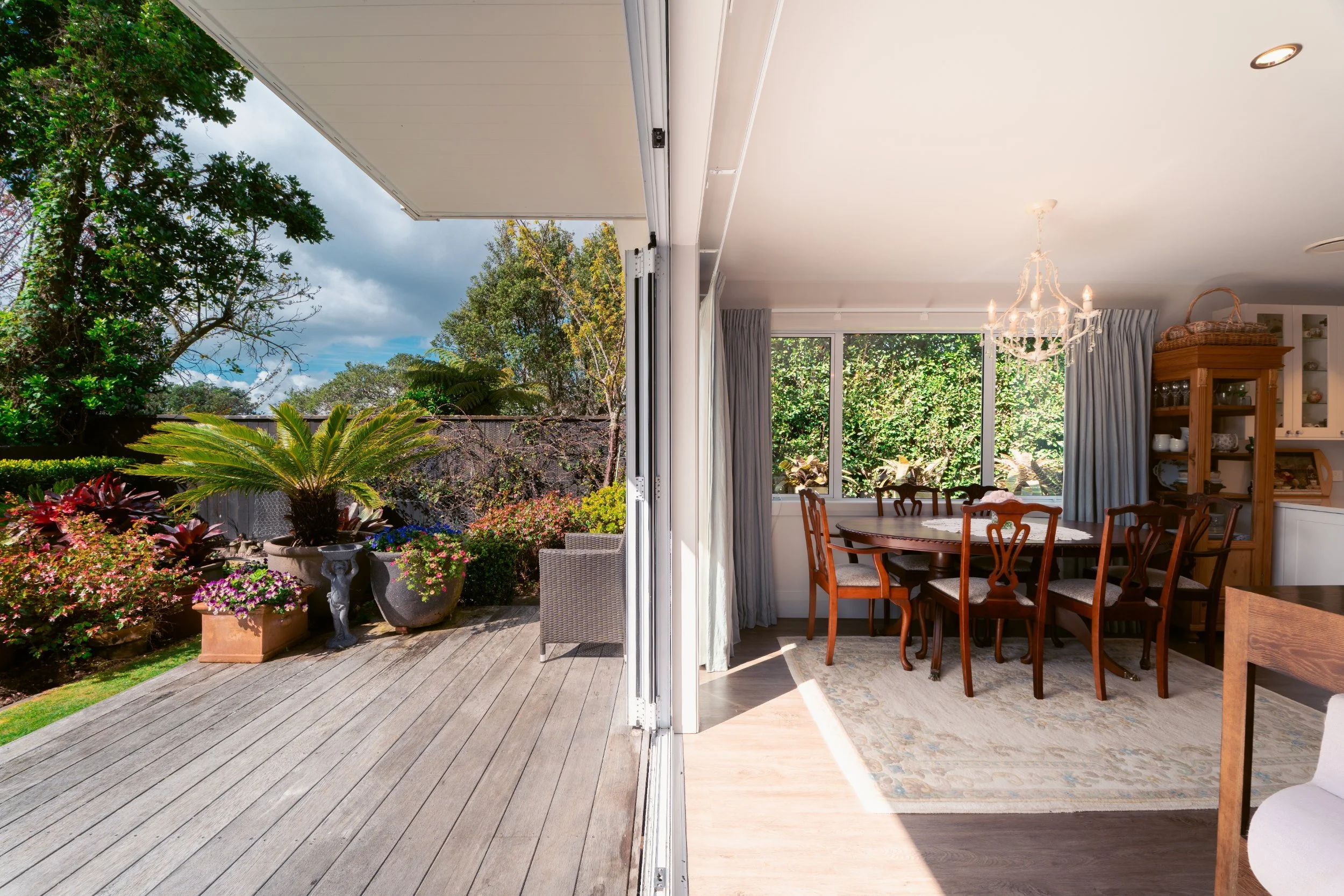 The image shows the interior of a dining room with wooden furniture including a round table and chairs. The room has large windows with curtains and a chandelier hanging from the ceiling. Outside, there is a patio garden with potted plants and trees,