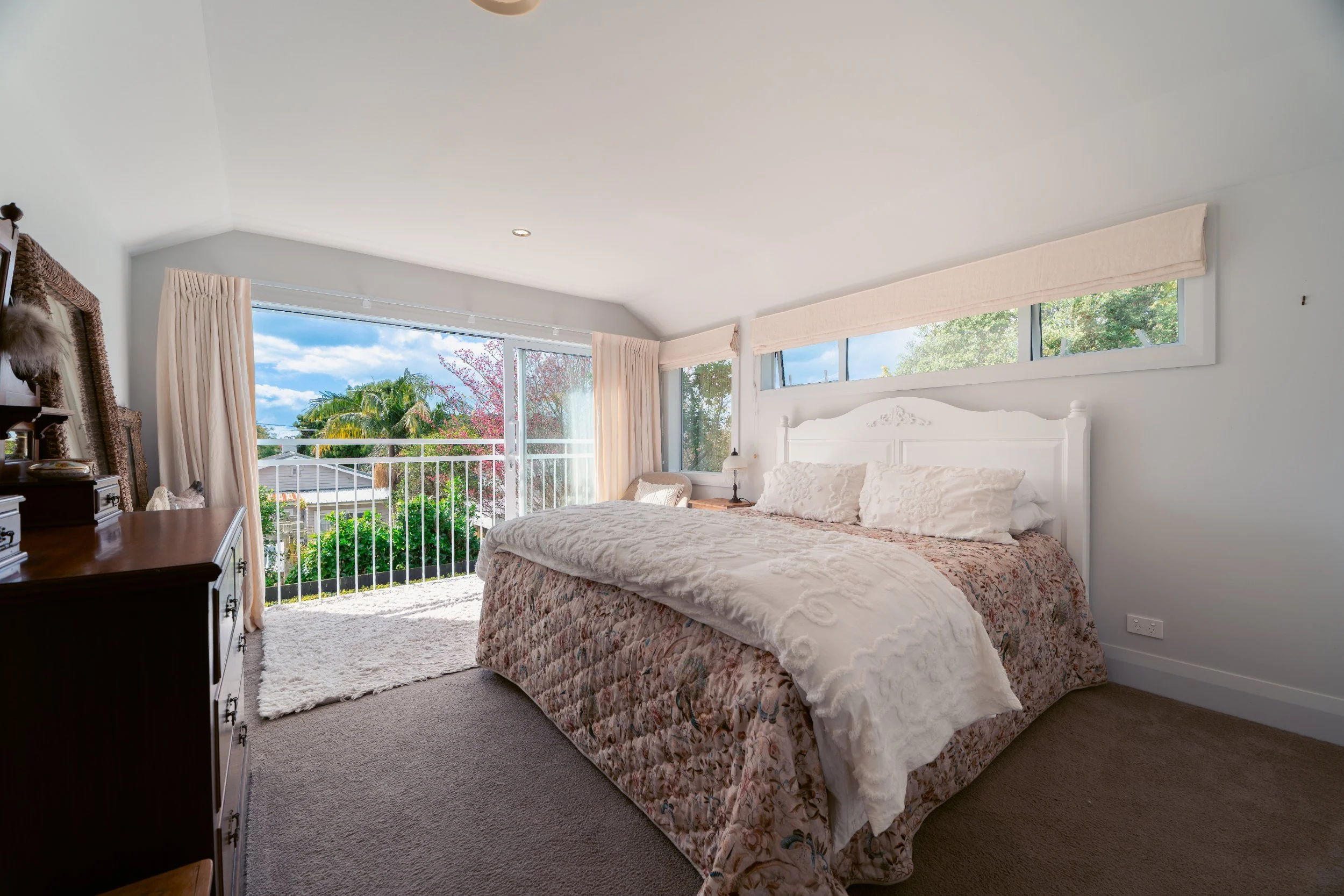 A bedroom with a large bed covered in floral bedding, a white headboard, and pillows. There's a window with a view of green trees and a balcony with a railing. Light curtains hang on the windows, and a dresser is partially visible on the left. The ro