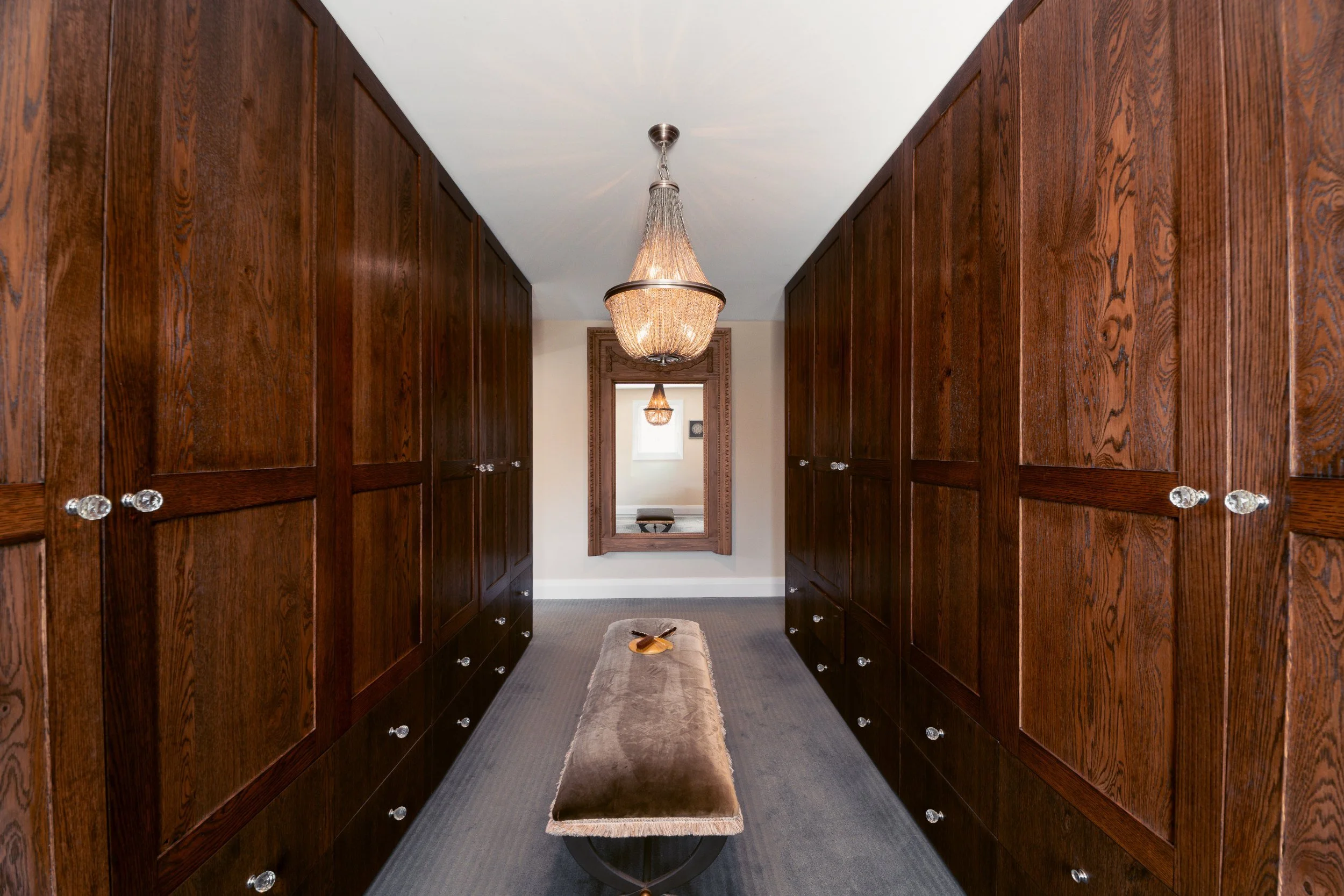 A walk-in closet with dark wooden cabinets on both sides, a beige carpet runner, and a chandelier hanging from the ceiling. A mirror is on the back wall.