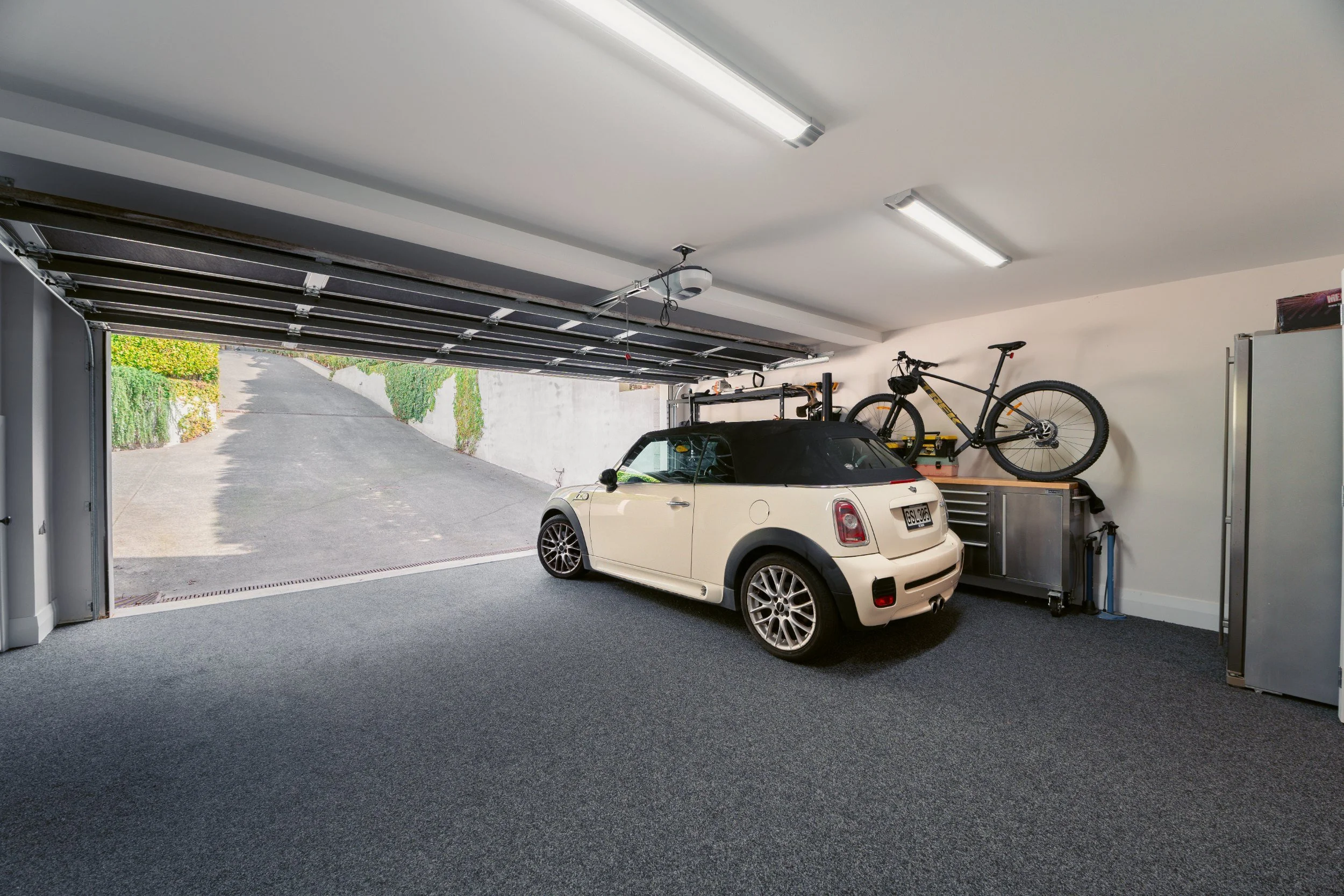 Inside a garage with an open door showing a driveway and greenery outside. Inside, there is a white Mini Cooper parked on a dark carpeted floor, with a bicycle mounted on a wall rack above it. The garage ceiling has bright fluorescent lights and a ga
