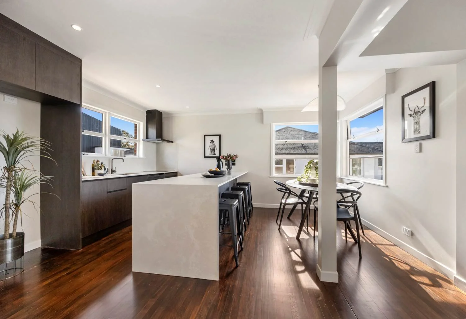 Modern kitchen with dark wood cabinetry, white countertops, a kitchen island with four black stools, a small round dining table with four chairs, and large windows with a view of neighboring houses and blue sky.
