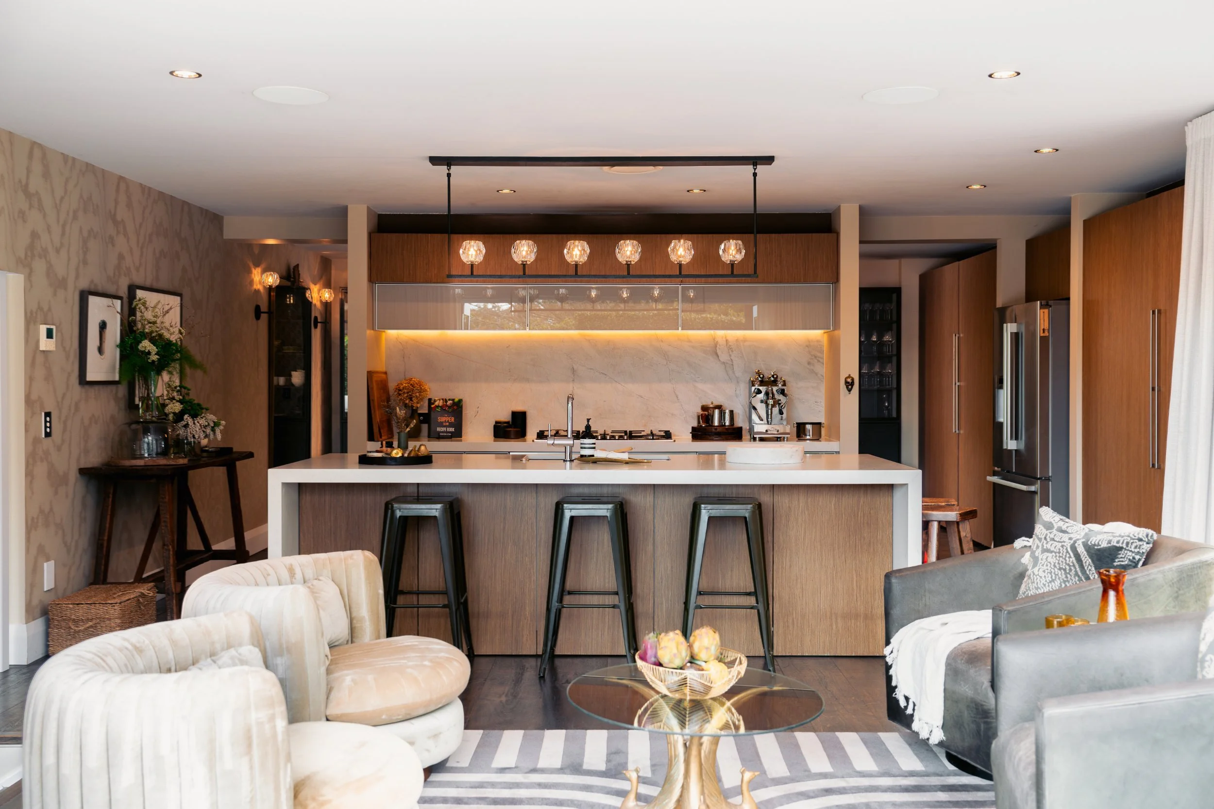 Modern kitchen with a white island, wood cabinets, pendant lighting, marble backsplash, and a cozy living area with beige and gray seating.