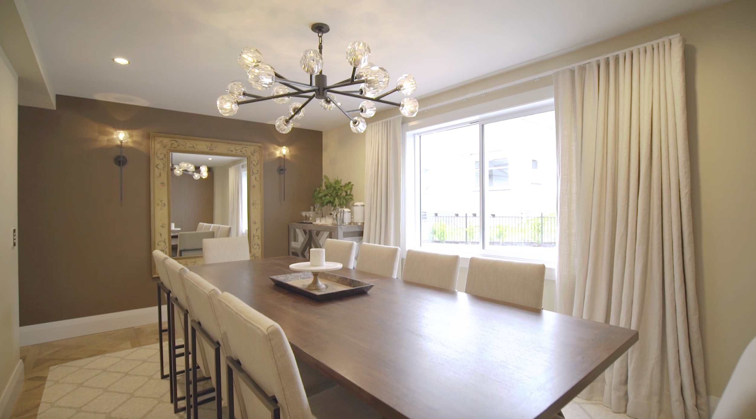 A modern dining room with a wooden table, cream-colored chairs, large window with beige curtains, a decorative mirror on the wall, and a contemporary chandelier.