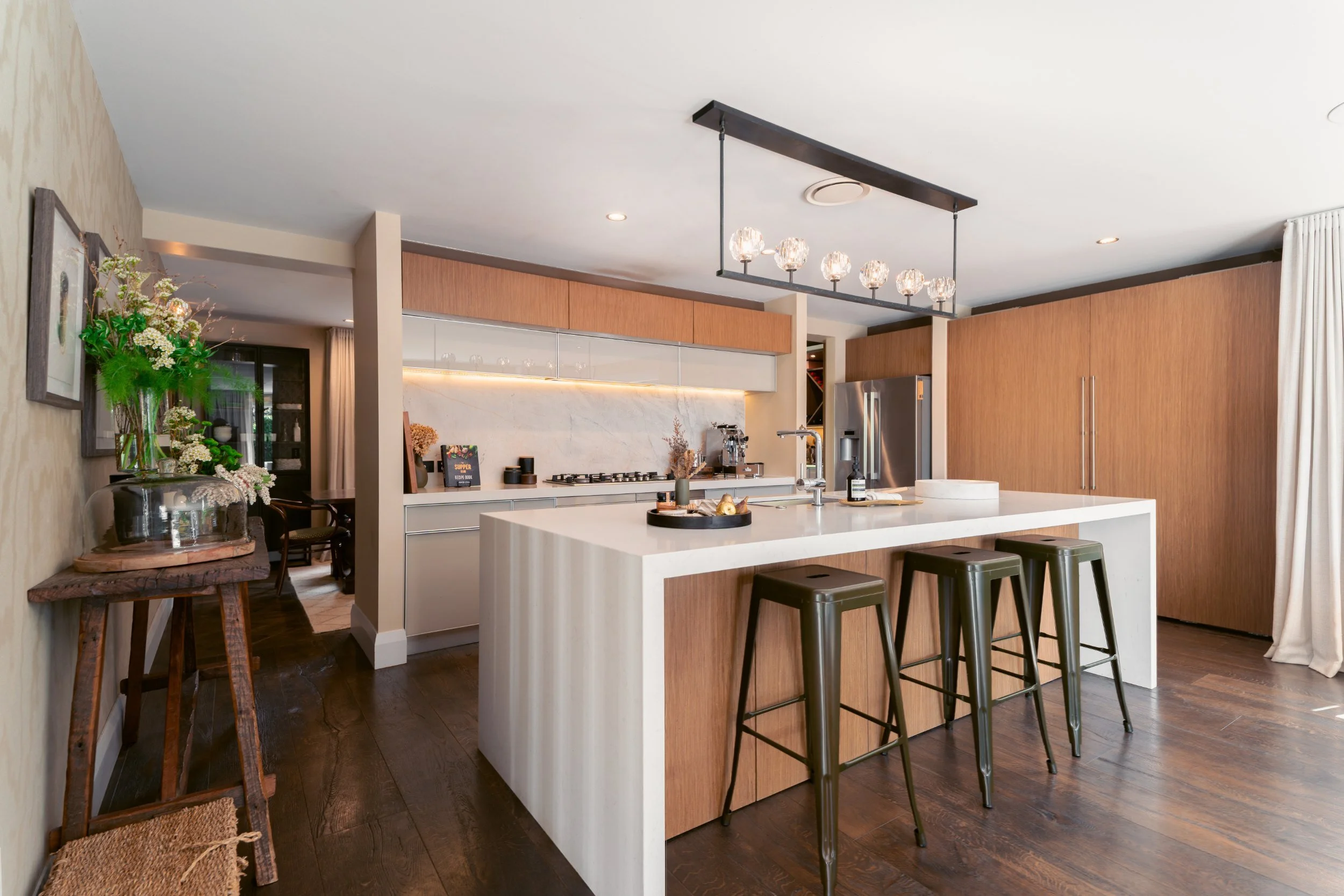 Modern kitchen with white island, wooden cabinets, stainless steel refrigerator, and black barstools; decorated with a vase of flowers and a tray of decor on the island.