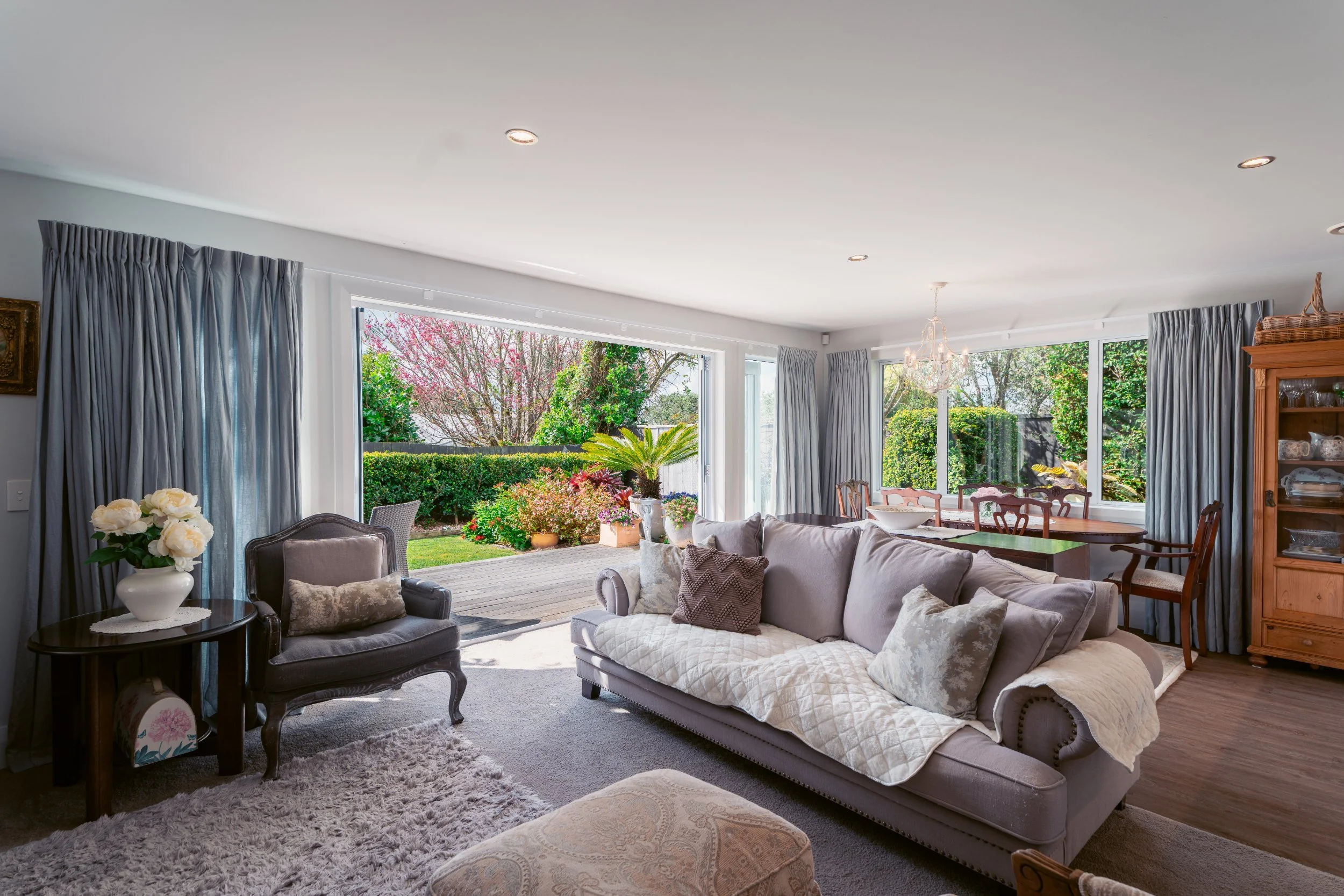 Living room with large windows showing a garden with trees and pink blossoms, furnished with sofas, armchairs, a wooden cabinet, and decorated with flowers and pillows, illuminated by natural daylight and a chandelier.