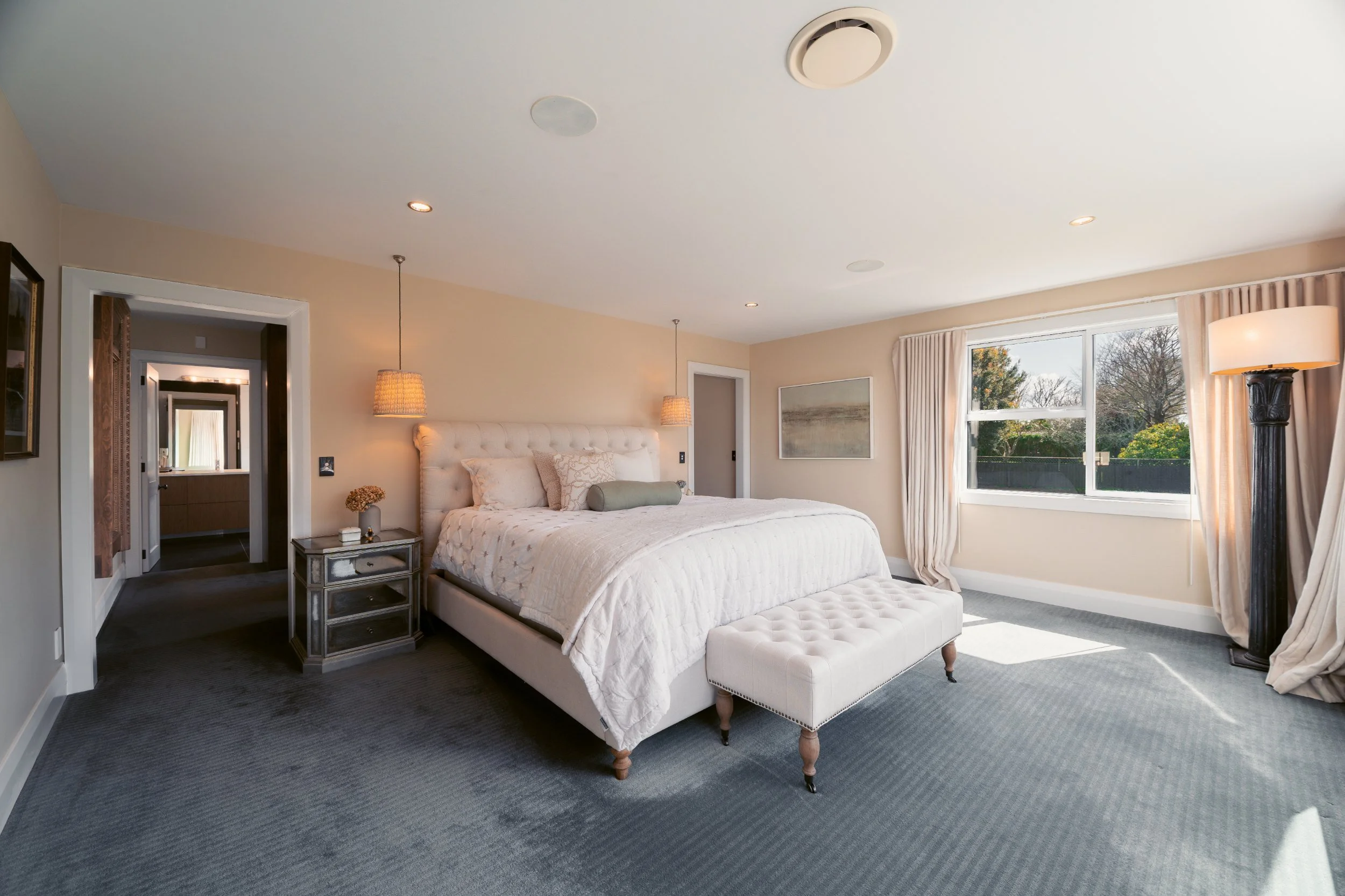 A bedroom with a large bed with white bedding, beige pillows, and a beige tufted headboard. There is a small mirrored nightstand with a lamp and a small decorative item. The room has a window with beige curtains and shows a view of trees outside. The