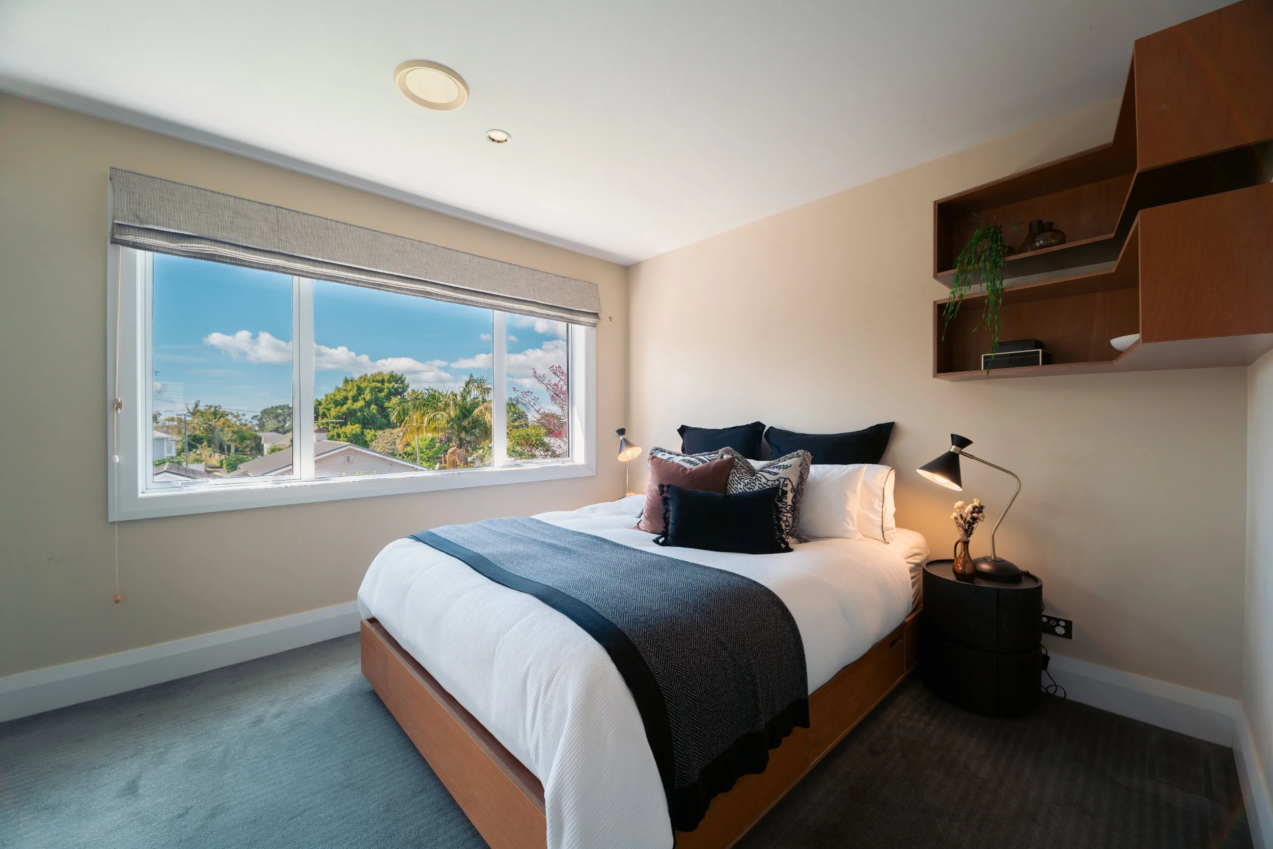 A bedroom with a large window showing trees and sky outside. The bed has white bedding with black and brown pillows. There are two black lamps on a round black nightstand. A wooden shelf adorns the wall above the bed.