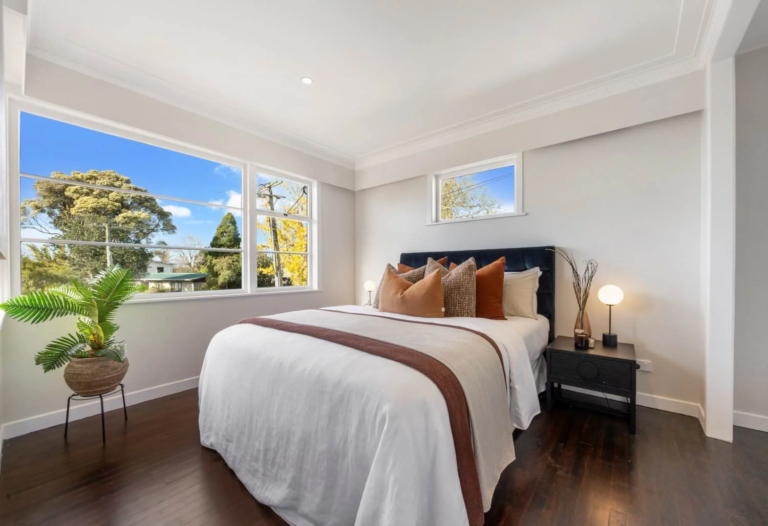 A bedroom featuring a bed with white bedspread and cushions, a black nightstand with a lamp and decorative items, a potted plant on a stand, large windows with an outside view of trees and utility poles, and hardwood flooring.