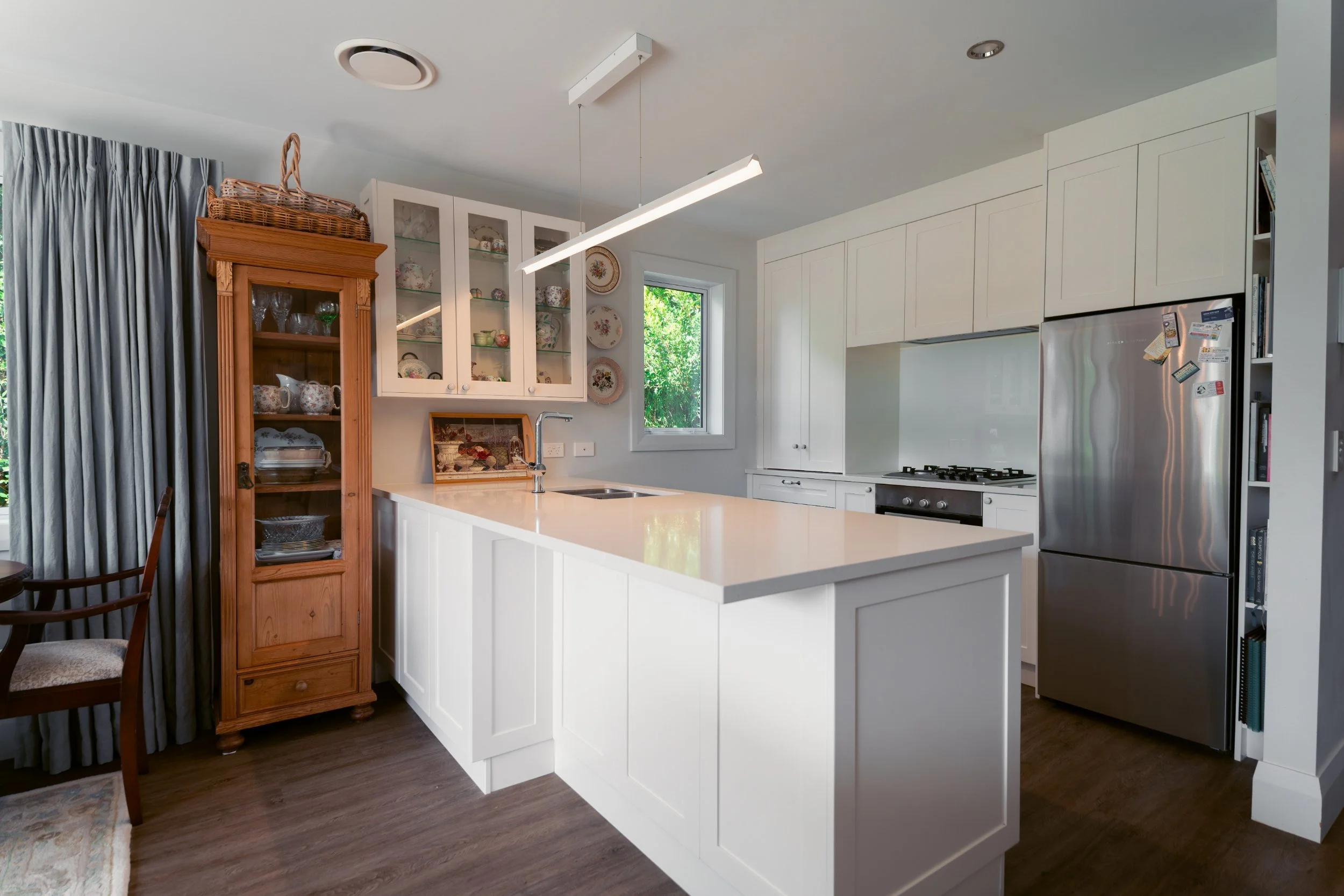 Modern white kitchen with island, stainless steel refrigerator, and glass-front cabinets filled with dishware.