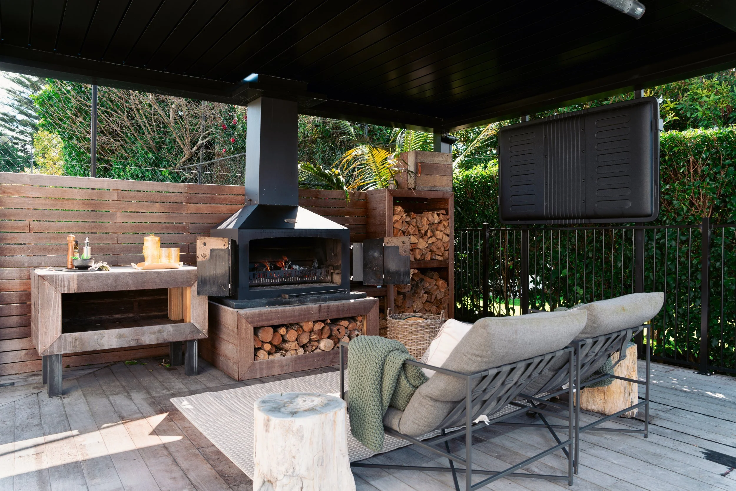 An outdoor patio area featuring a wood-burning fireplace, a stack of firewood, a wooden side table with candles and kitchen supplies, seating with cushions, and lush green foliage in the background.