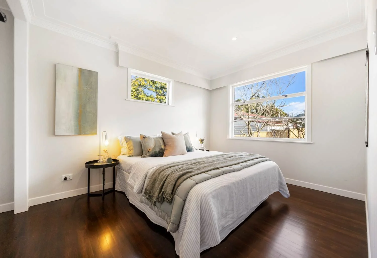 Bright bedroom with white walls, a bed with white and beige bedding and multiple pillows, a small round black side table with a lamp and decor, large windows showing trees outside, wooden flooring, and minimal wall art.