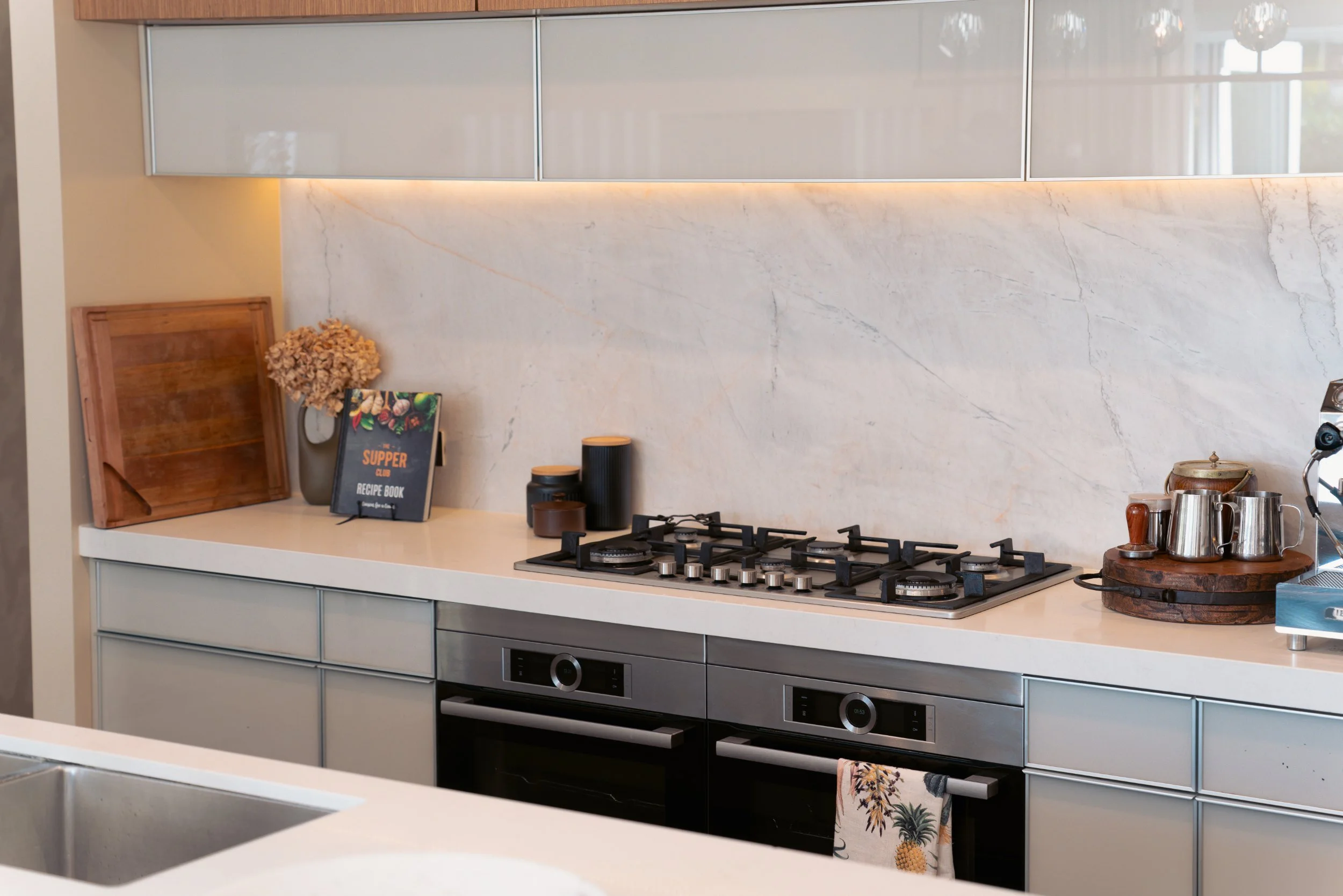 Modern kitchen with marble backsplash, gray cabinets with frosted glass, a gas stove, a wooden tray, a cookbook titled 'Supper Club Recipe Book,' a vase with dried flowers, and various kitchen utensils on the countertop.