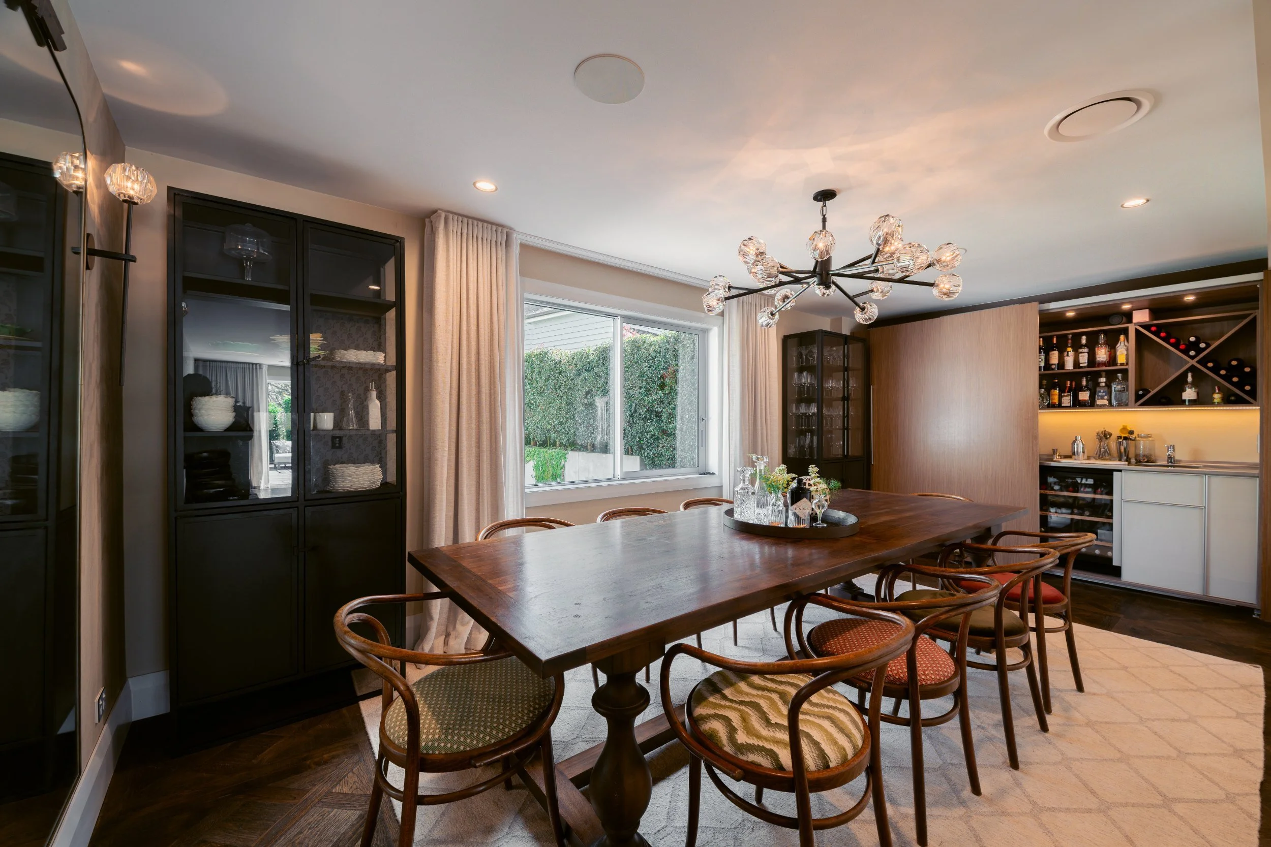 Interior of a dining room with a large wooden table, multiple vintage-style chairs with patterned cushions, black display cabinets, a window with curtains, and a modern chandelier with glass globes.