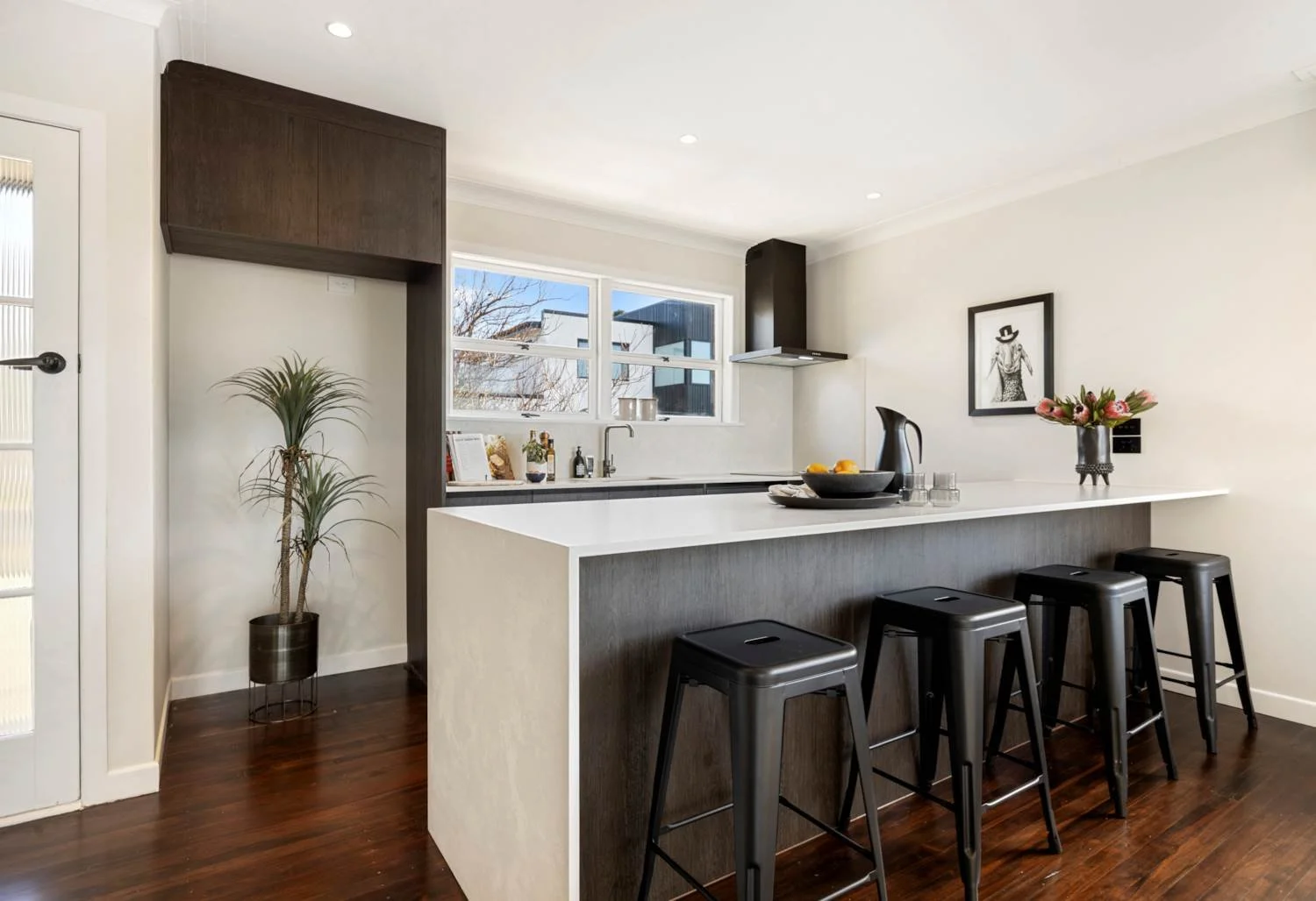 Modern kitchen with white and dark wood cabinetry, black bar stools, a flower vase, and a window view of neighboring buildings.