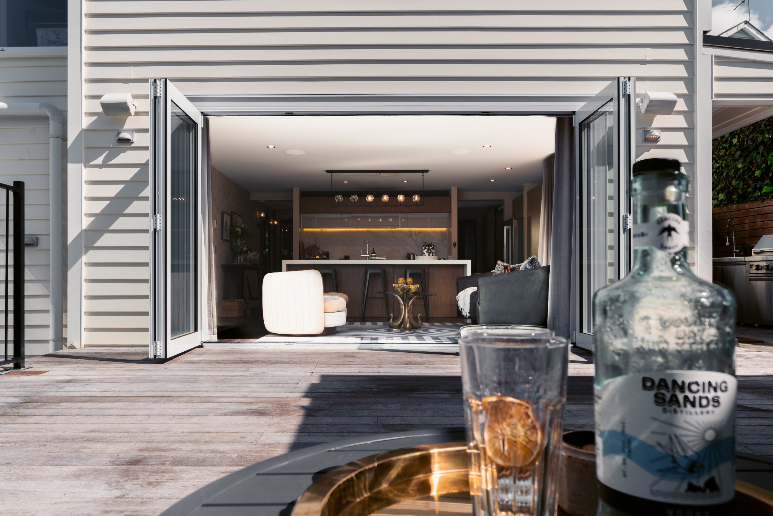 Open patio door revealing a modern indoor living space with a kitchen and seating area, seen from an outdoor deck with wine glasses, a bottle of sand dunes flavored snack, and a decorative tray in the foreground.