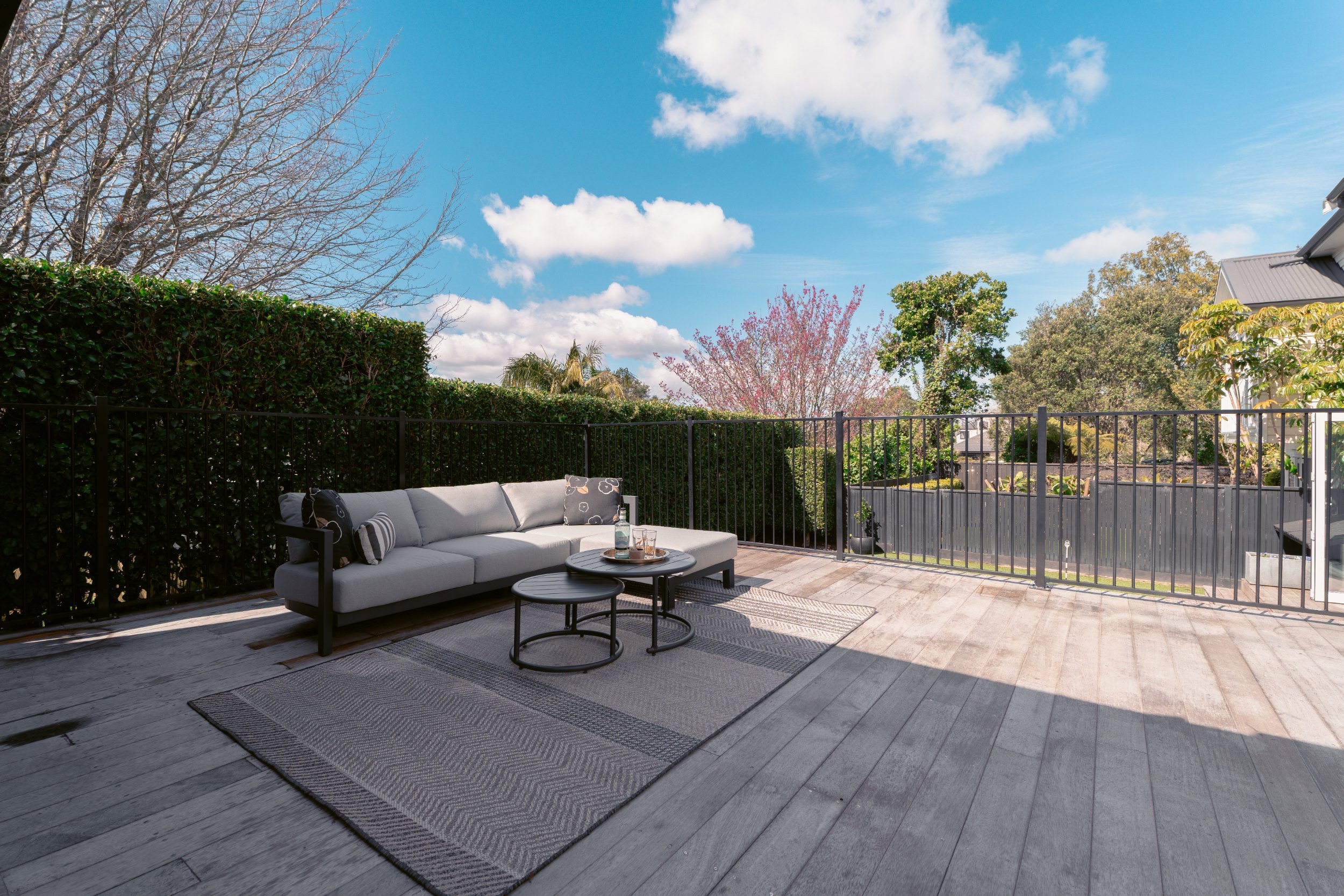 Outdoor patio with grey cushioned sofa, two black round tables, and patterned rug. Surrounding trees and bushes, blue sky with white clouds, and neighboring houses visible.