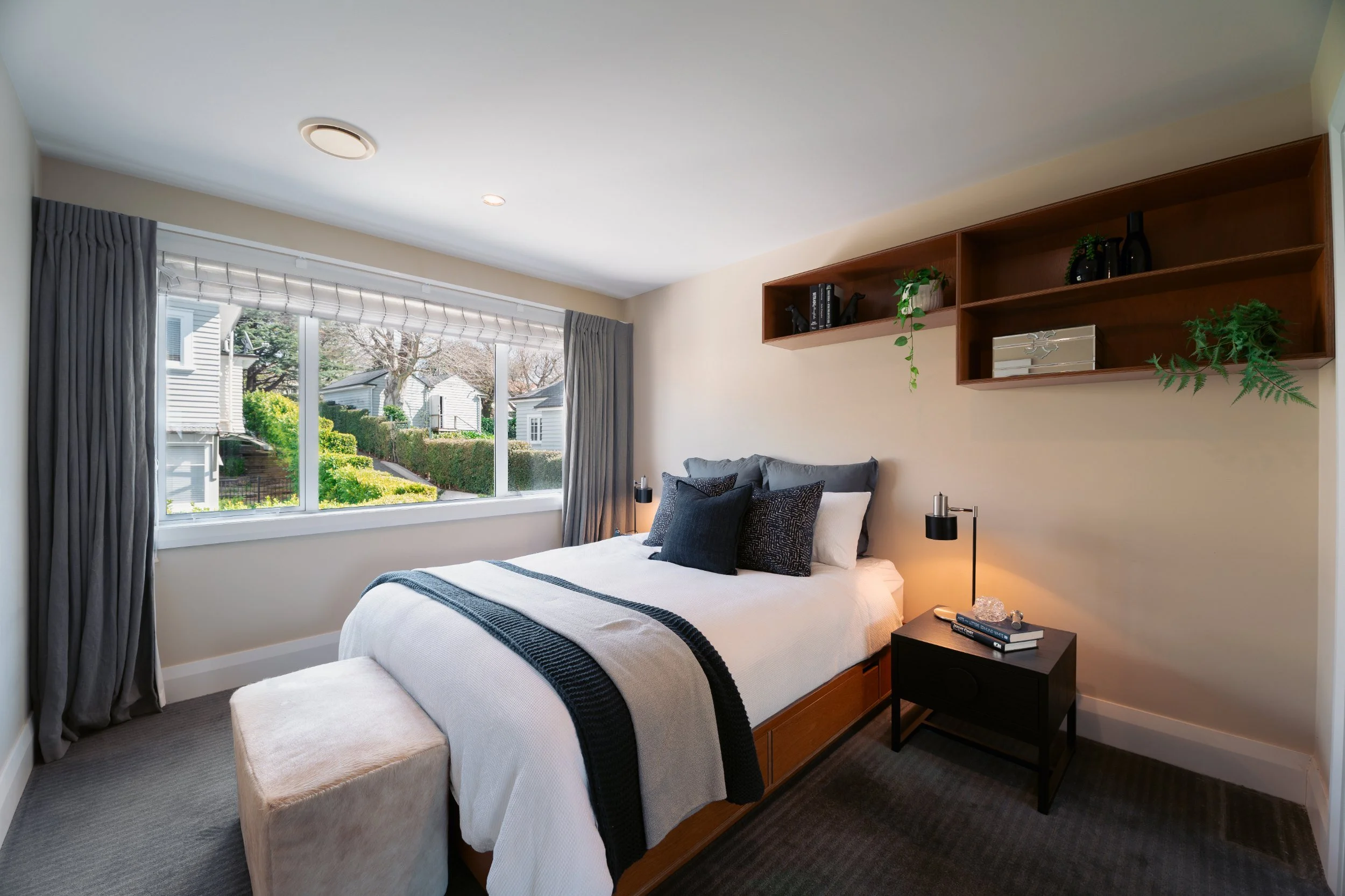 A cozy bedroom with a large window showing a view of neighboring houses and trees. The bed has dark pillows and a blanket, a small black bedside table with books and a lamp, and a wooden shelf with decorative items and plants mounted on the wall.
