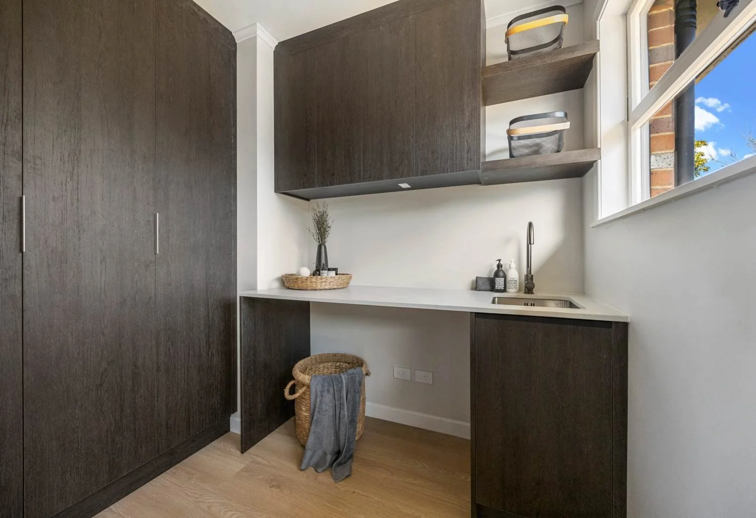 A minimalist laundry room with dark wood cabinets, a white countertop, a small sink, and a window showing a blue sky with clouds. Decorative items include two bottles of soap, a wicker basket with a gray towel, and small storage baskets on open shelv