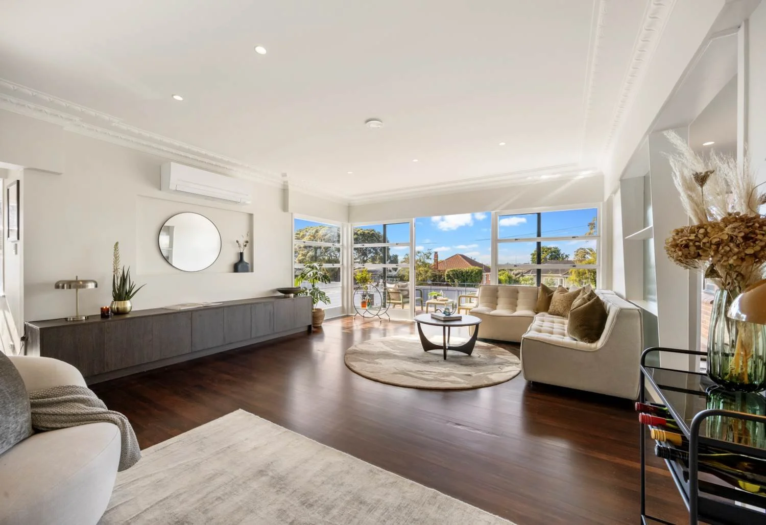 Bright living room with large windows, white ceiling and walls, dark wooden floor, round area rug, beige sectional sofa, coffee table, side table with plants and decor, and outdoor patio with chairs visible through windows.
