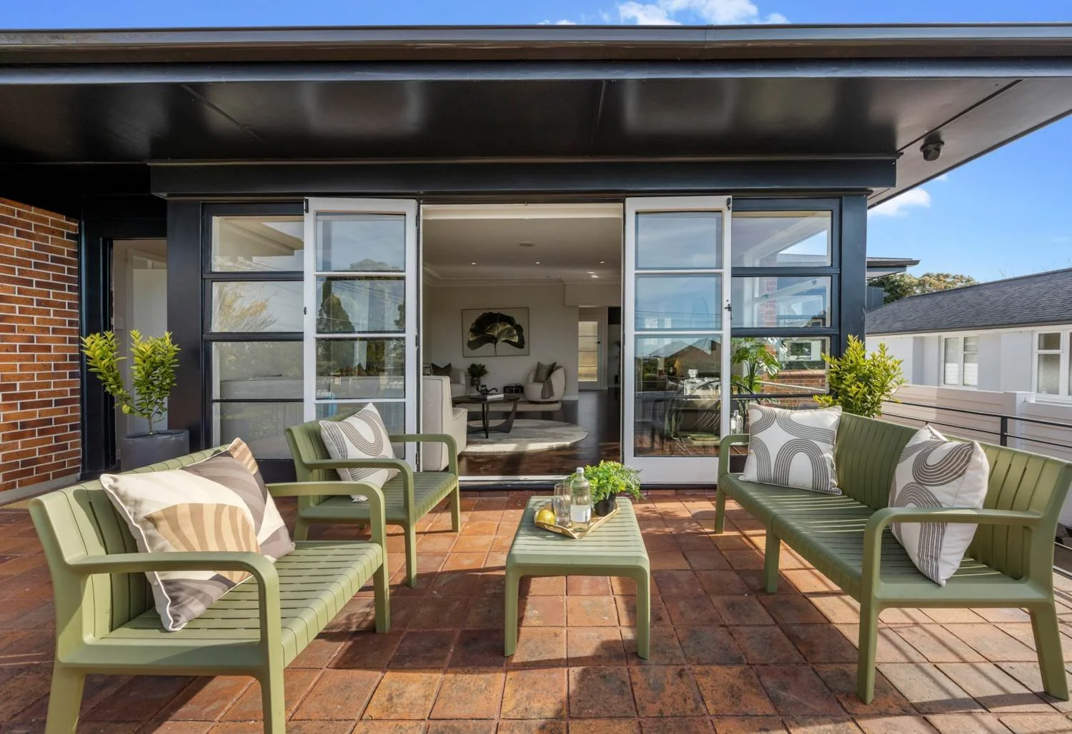Outdoor patio with green seating, pillows, plants, and view of the interior living room through glass doors
