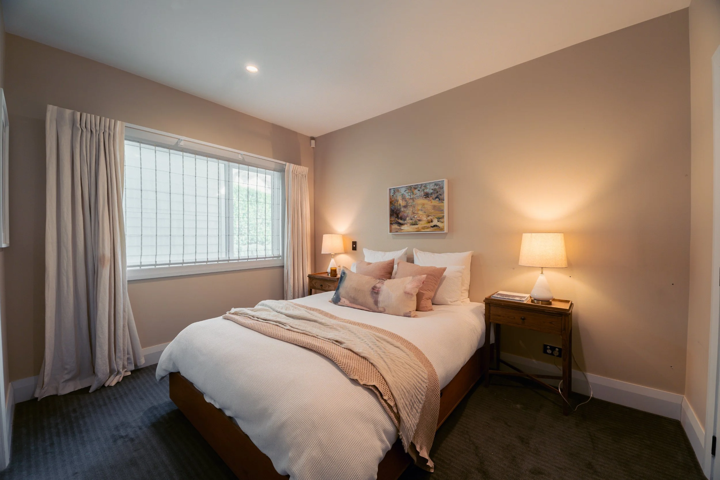 A cozy bedroom with a large window, beige walls, a bed with white bedding and multiple pillows, wooden nightstands with lamps, and a framed landscape painting on the wall.