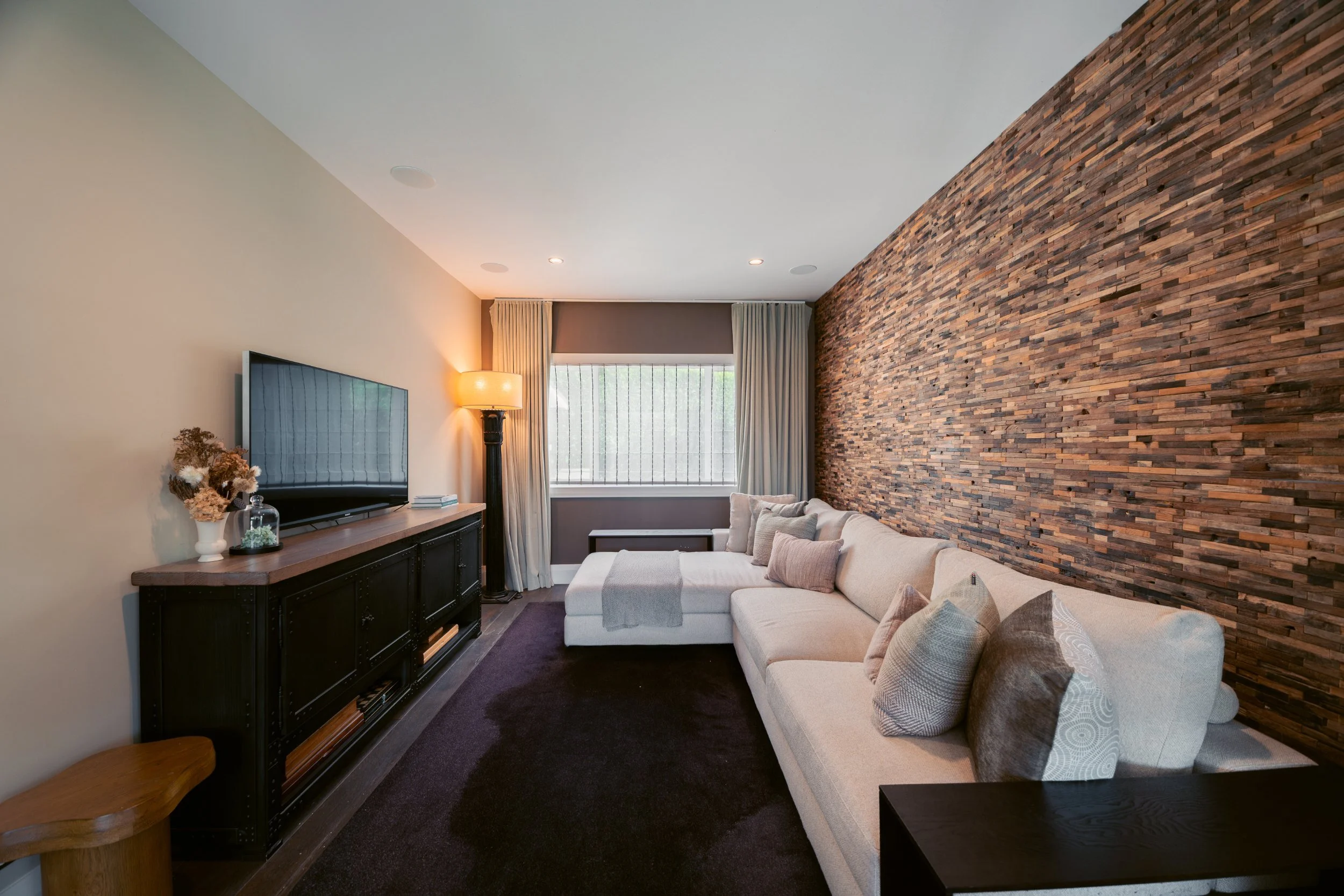 Living room with a white sectional sofa, a black console table, a large flat-screen TV, a tall black floor lamp, beige curtains, a window, a brown brick accent wall, and decorative pillows.