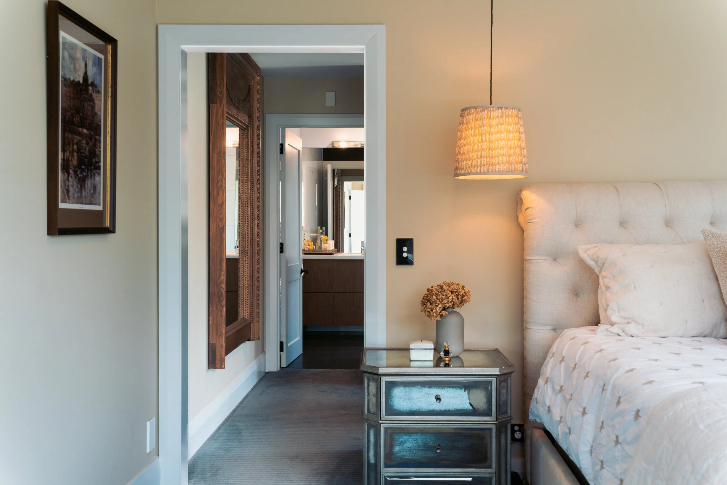 A bedroom with a beige upholstered bed, a silver mirrored dresser, a window with a wooden frame, a bedside lamp, a framed painting on the wall, a small flower vase with dried flowers, and an open door leading to a bathroom with a sink and mirror.
