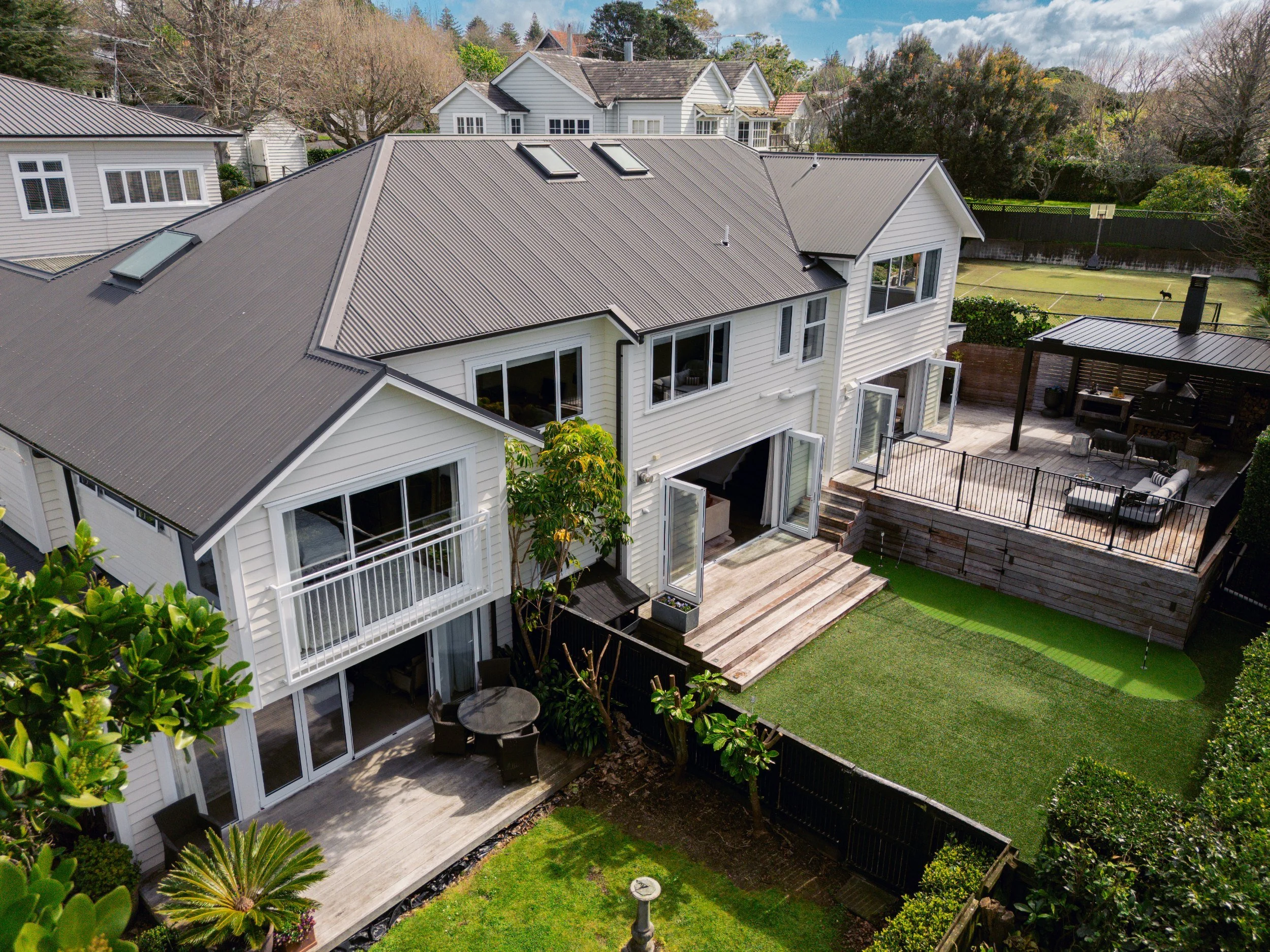 Aerial view of a residential backyard with a wooden deck, patio furniture, a grassy lawn, a basketball hoop, and neighboring houses with similar architectural styles and roofing.