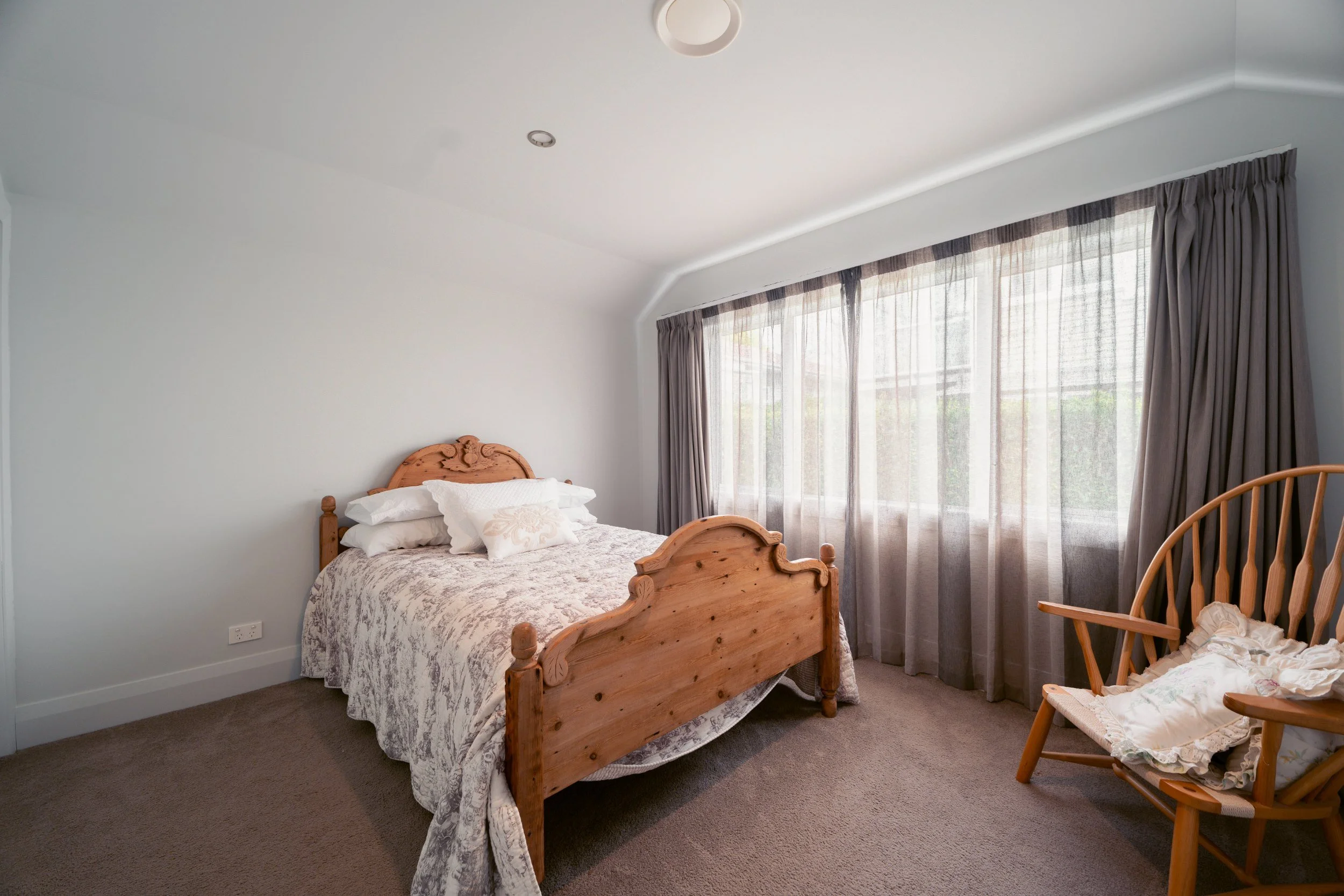 A bedroom with a wooden bed, white bedding, and pillows. Large window with sheer curtains and gray drapes, brown carpet, and a wooden rocking chair with blankets on it.